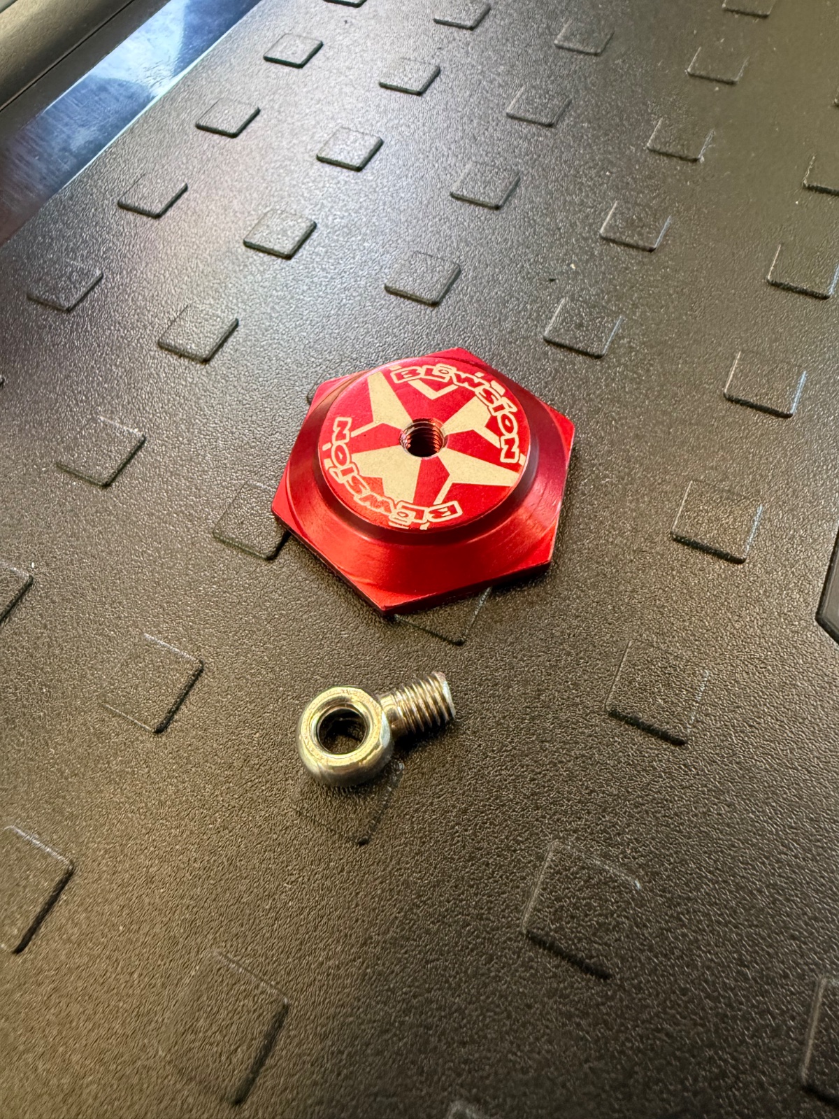 Red anodized aluminum cap with cross-pattern design and brass threaded insert displayed on textured rubber mat with a small brass eyelet below it