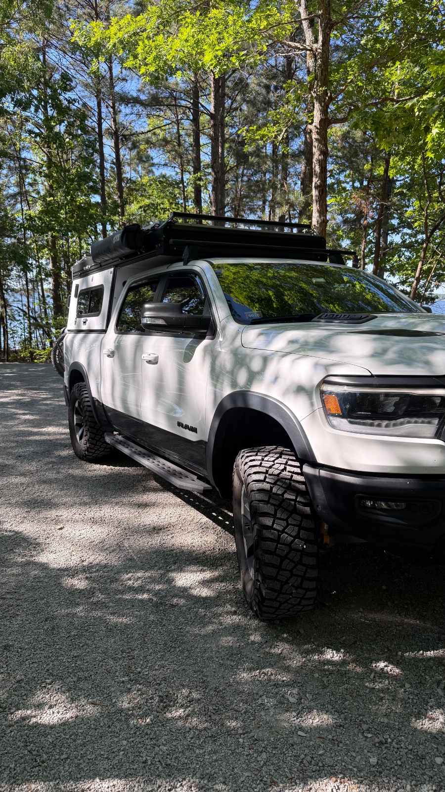 White Dodge Ram truck with roof rack and all-terrain tires parked on gravel under tall pine trees on a sunny day