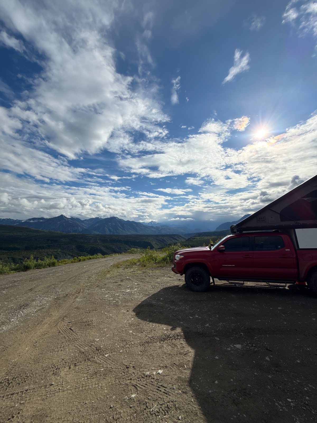 Red Toyota Tacoma with roof rack parked on gravel mountain overlook with vast forested valley, distant snow-capped peaks, and dramatic blue sky with white clouds