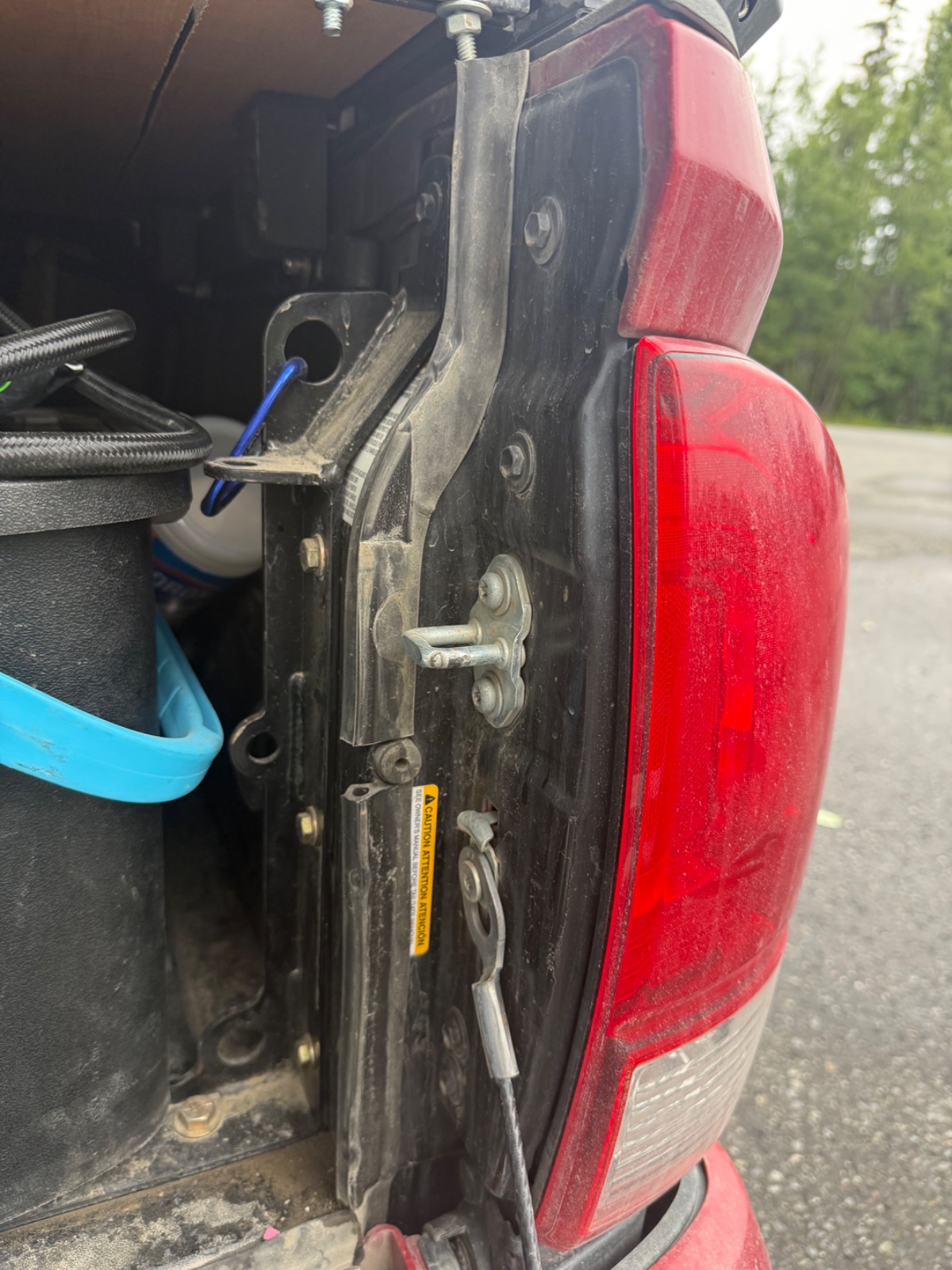Close-up of a vehicle's open rear door showing red taillight assembly, black interior panel, blue wiring, and metal hinges on a paved road with green trees in background