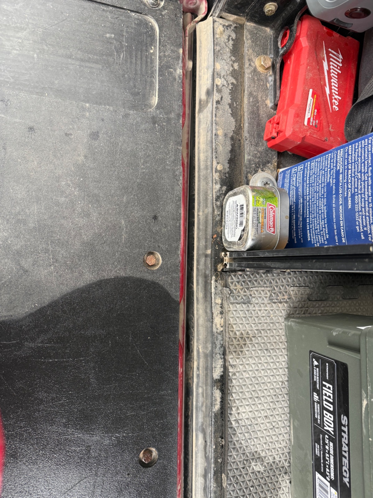 Truck bed view showing Milwaukee tool bag, Coleman cooler, blue instruction manual, and diamond plate toolbox with metal cleats visible on overlanding vehicle