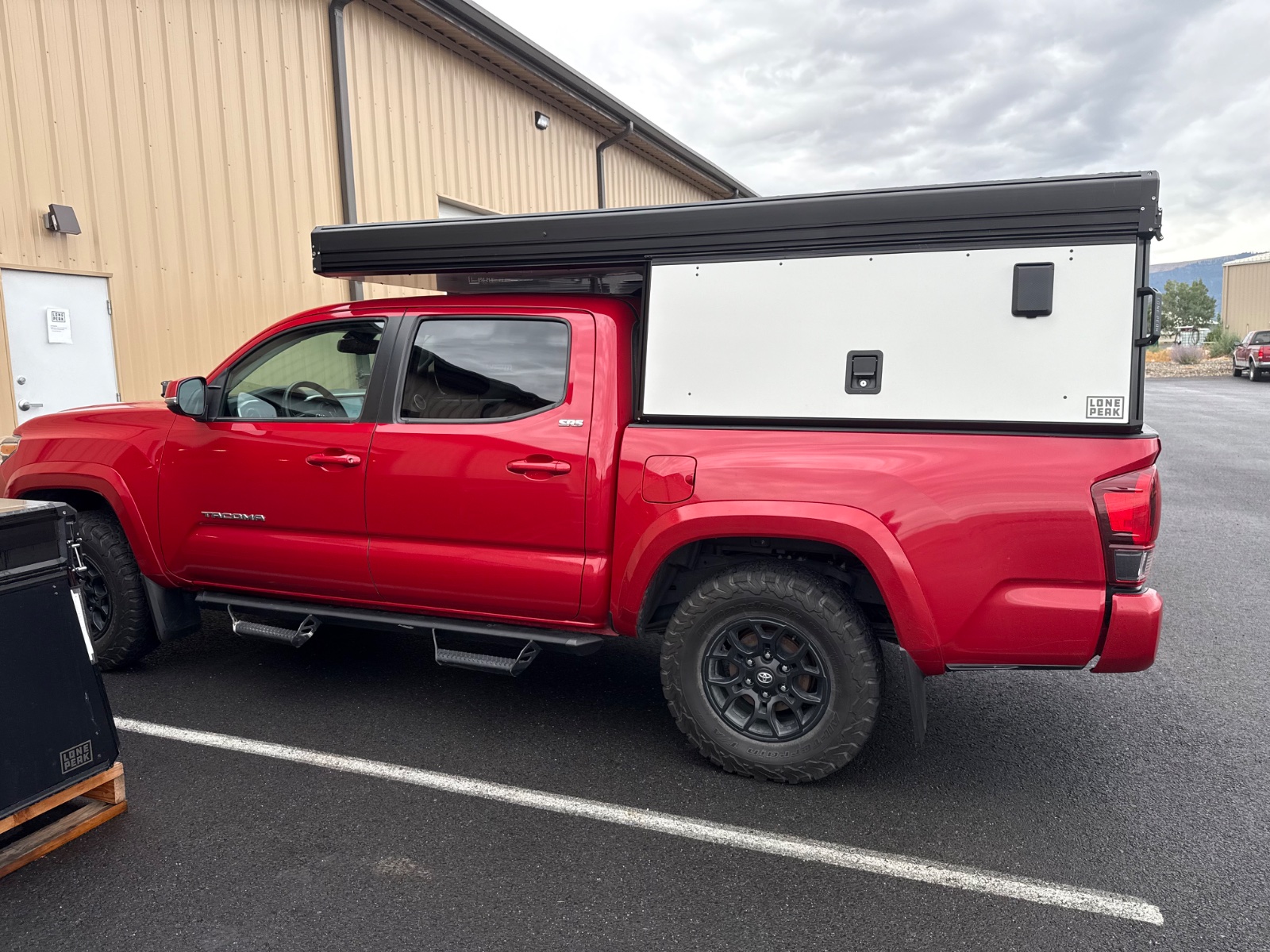 Red Toyota Tacoma pickup truck with white Lone Peak camper shell in a parking lot near industrial buildings under cloudy skies