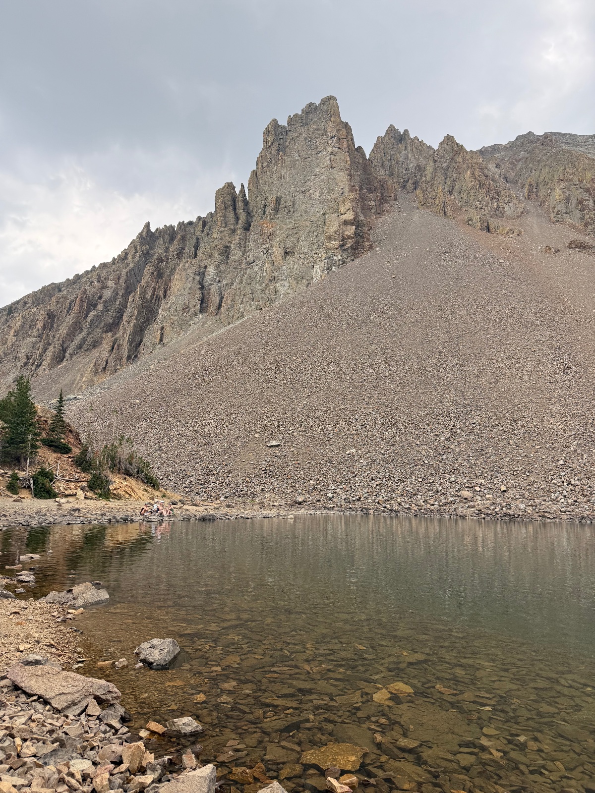 Alpine lake surrounded by rocky scree slope and dramatic columnar basalt rock formations under overcast sky