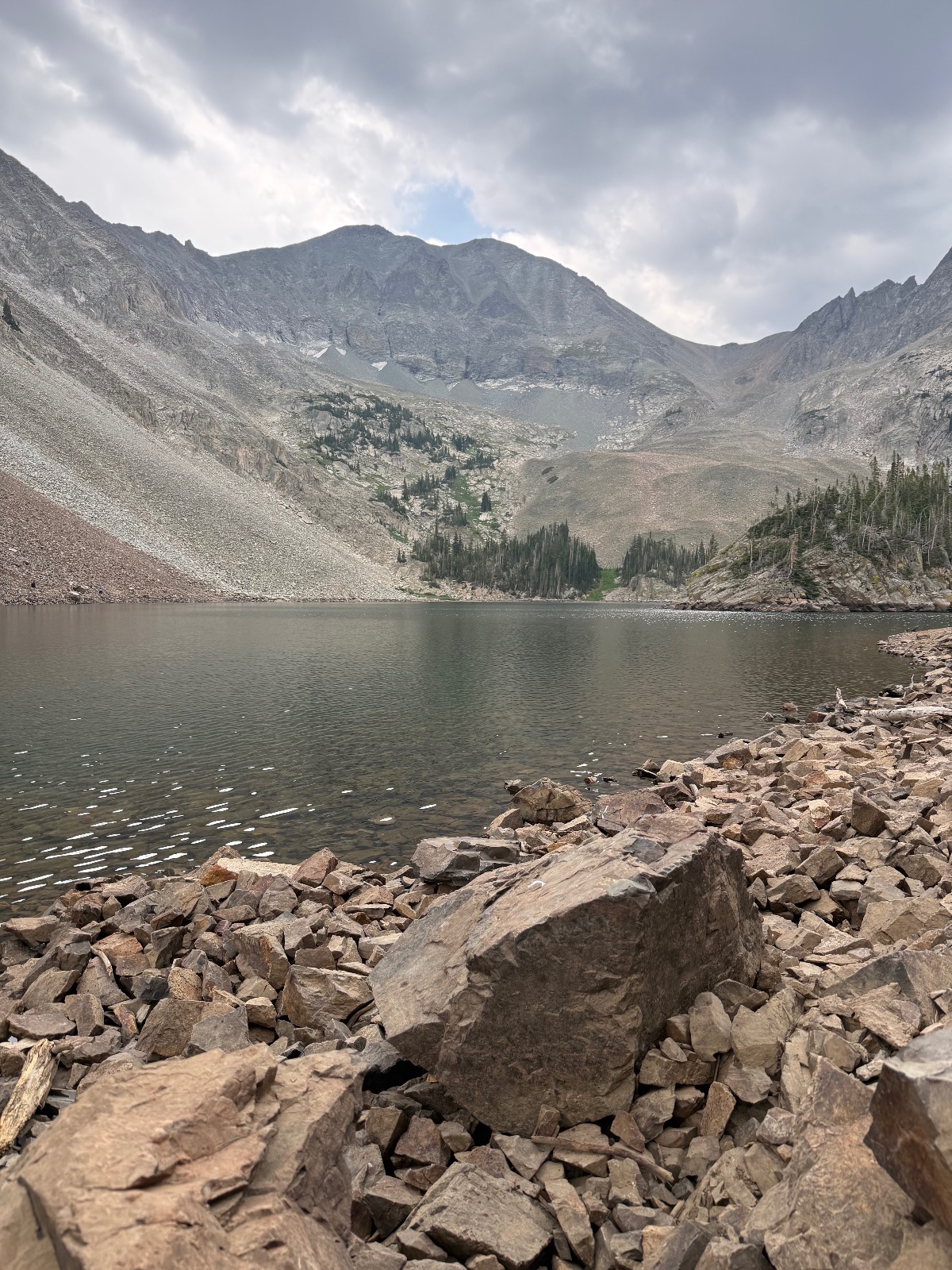 Rocky shoreline of an alpine lake surrounded by evergreen forests and bare mountain peaks under cloudy skies