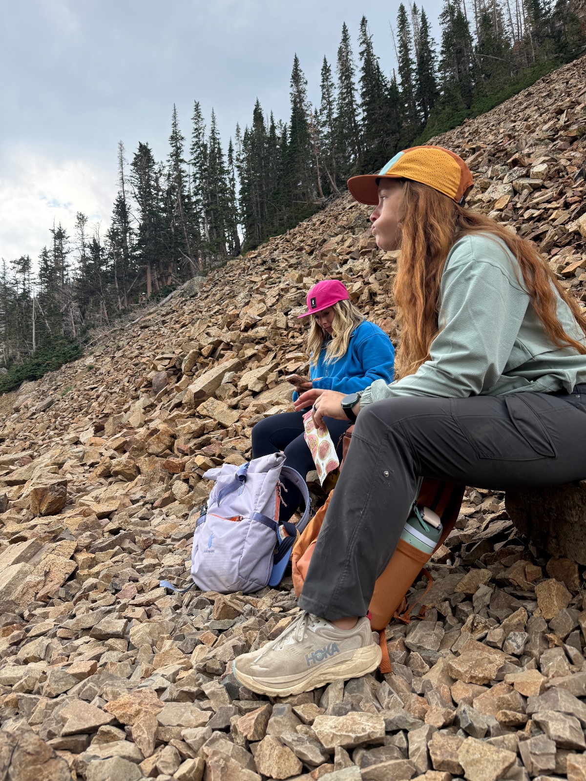 Two hikers in colorful caps rest on a rocky scree slope with coniferous forest and overcast sky in background