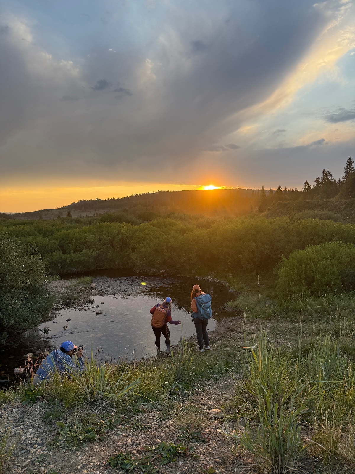 Three hikers stand by a stream at sunset with forested mountains and golden sky in the background during evening trek