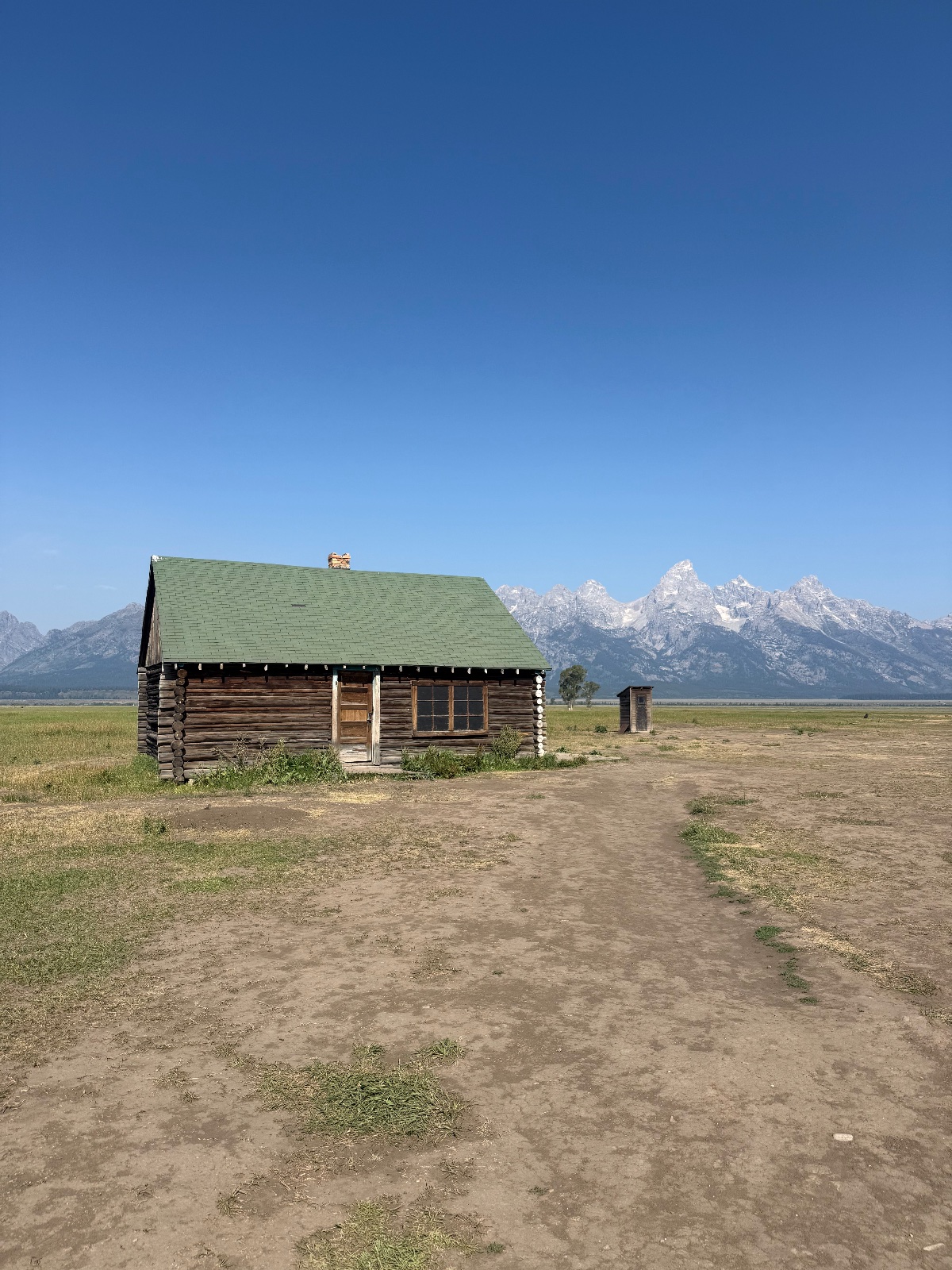 Historic log cabin with green roof on flat meadow valley with snow-capped mountain range backdrop under clear blue sky