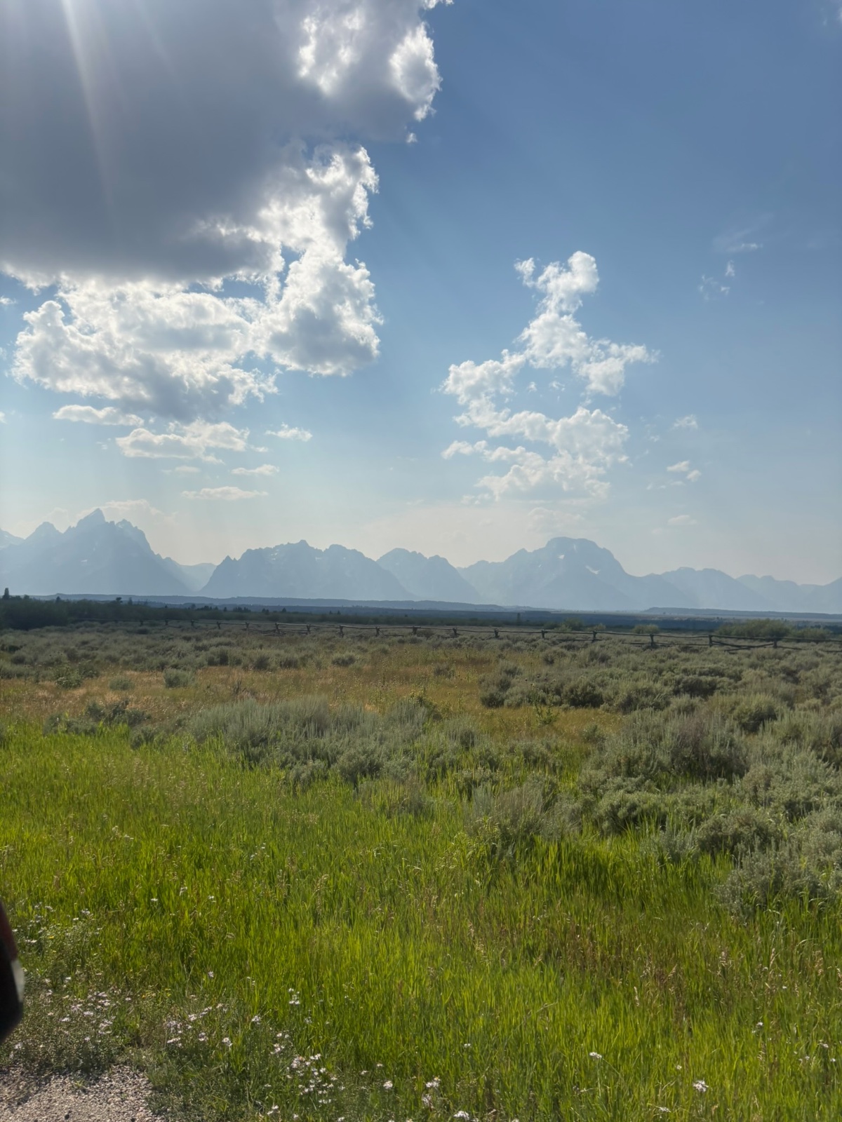 Wide valley landscape with sagebrush vegetation, distant jagged mountain range, scattered fence posts, and puffy white clouds under blue sky