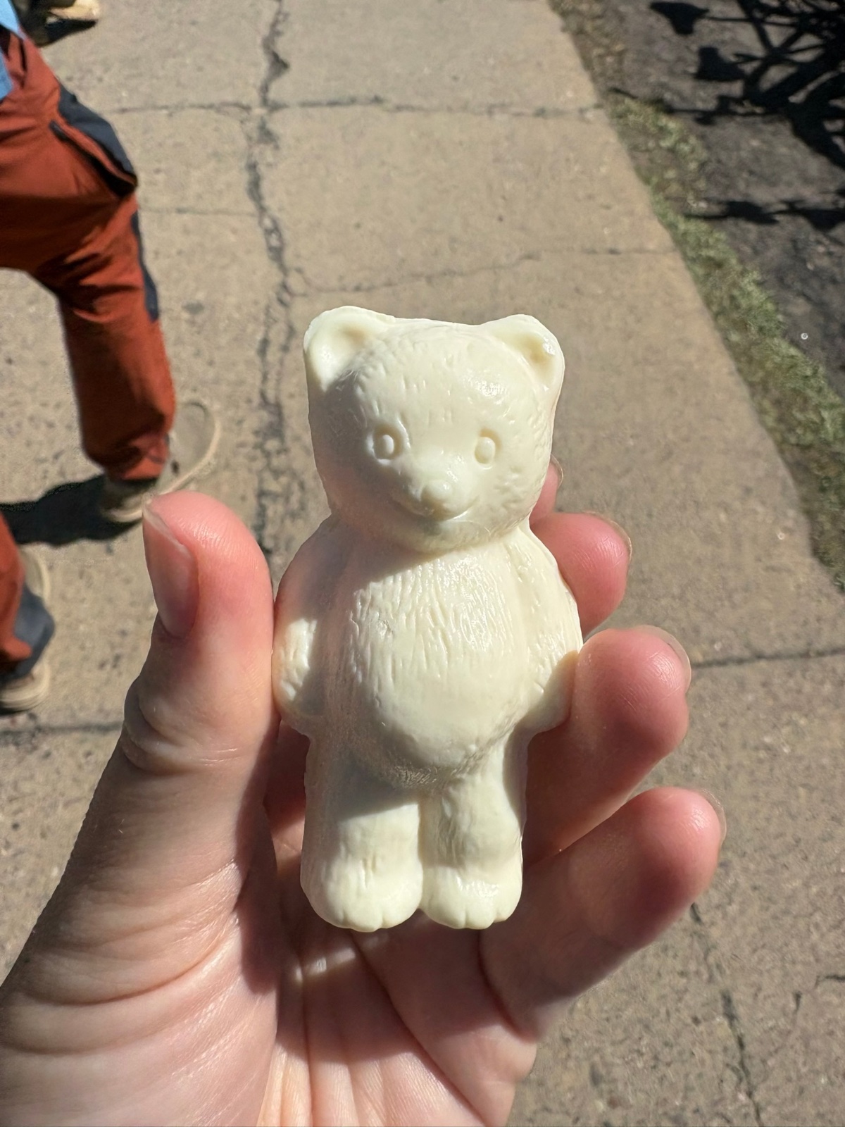 A small white teddy bear figurine held in someone's hand on a paved outdoor path