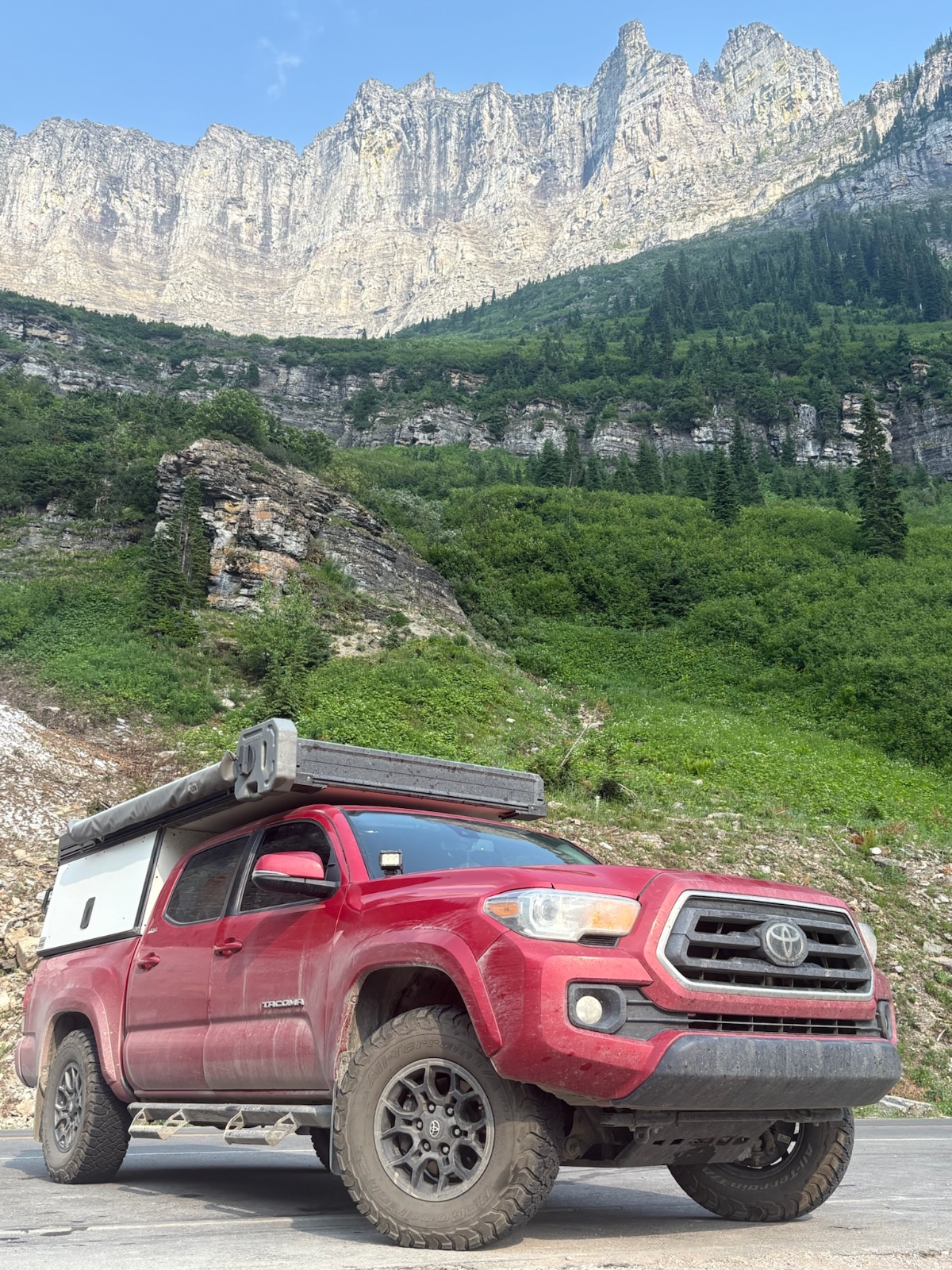 Red Toyota Tacoma with roof rack parked on gravel overlook facing massive columnar basalt cliffs and forested valley under clear blue sky