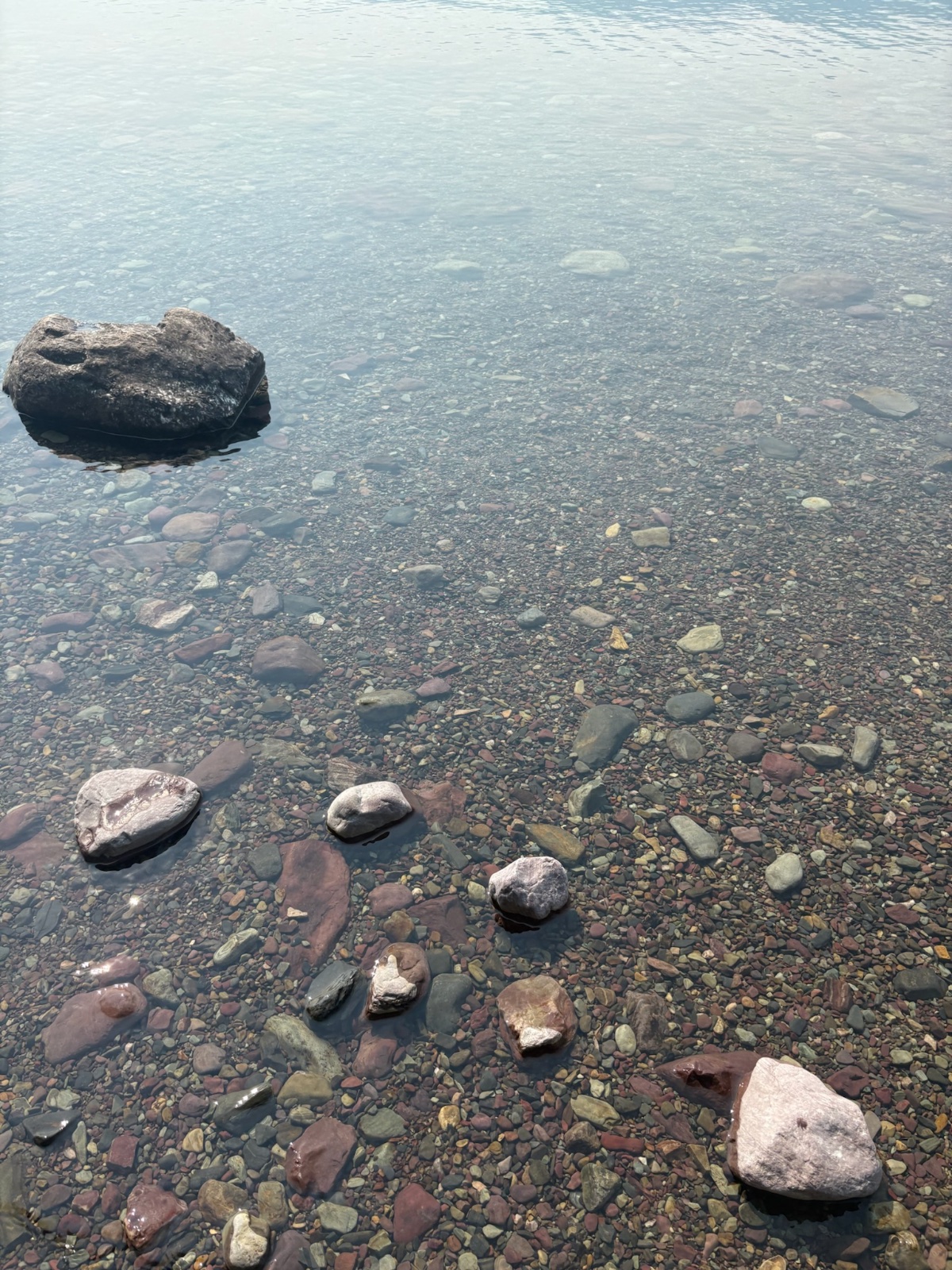Clear shallow water with visible rocks and pebbles on a beach, featuring a large boulder in the upper left and smooth stones scattered across the sandy bottom