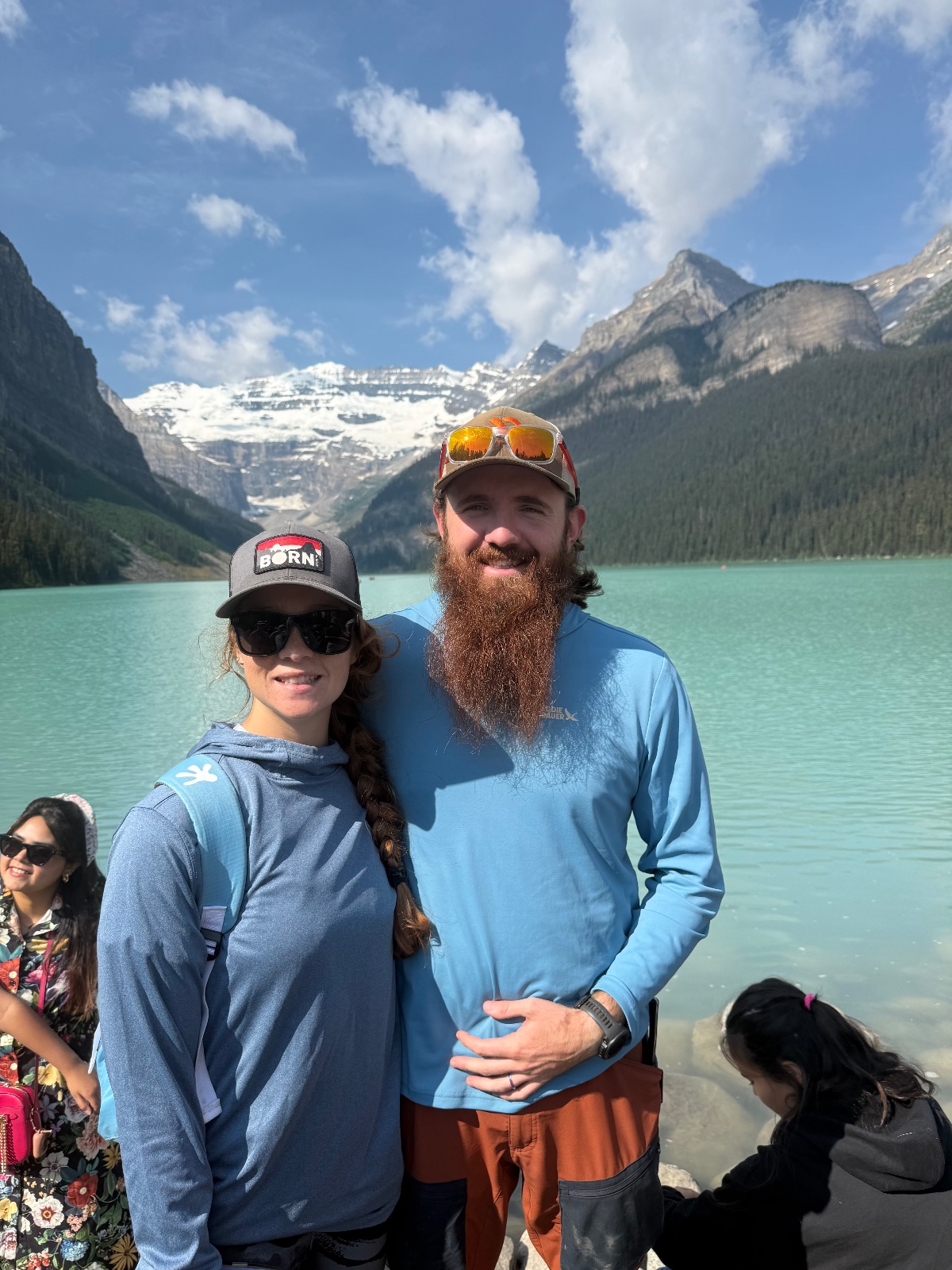 Two people smiling at turquoise alpine lake with snow-capped mountains and evergreen forest backdrop under blue sky