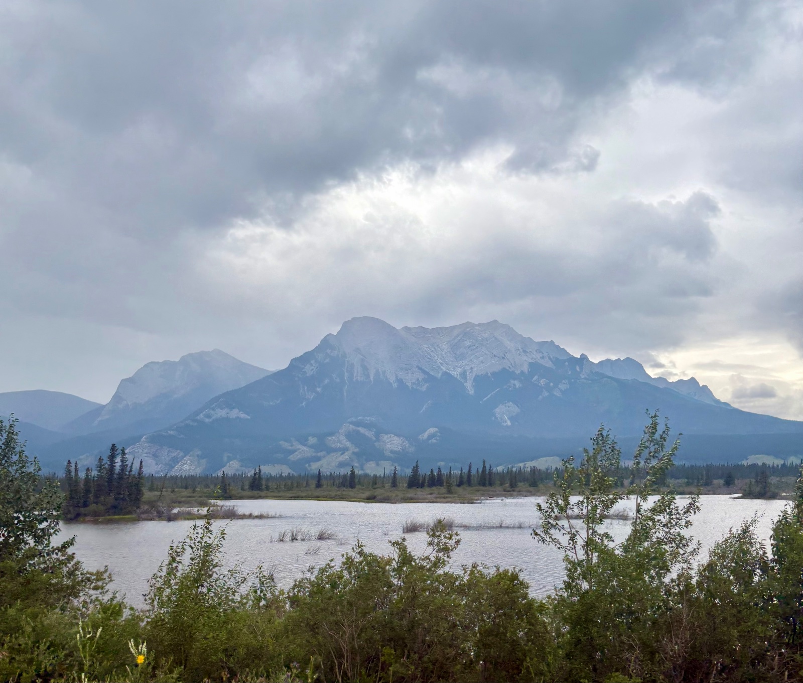 Snow-capped mountain rising above a forested shoreline and calm lake under dramatic cloudy sky in boreal landscape