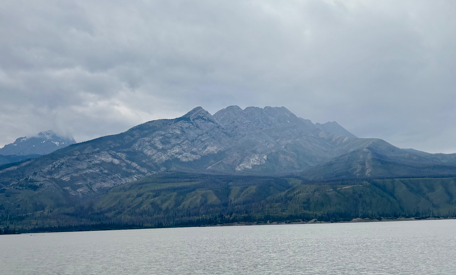 Mountain lake view with rocky peaks and dense conifer forest under overcast sky, glacial water shoreline visible