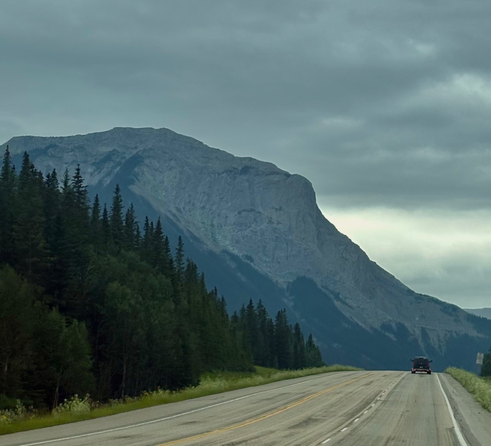 Straight highway stretches toward a dramatic rocky mountain peak with dense coniferous forest on both sides under cloudy skies, with a truck visible in the distance