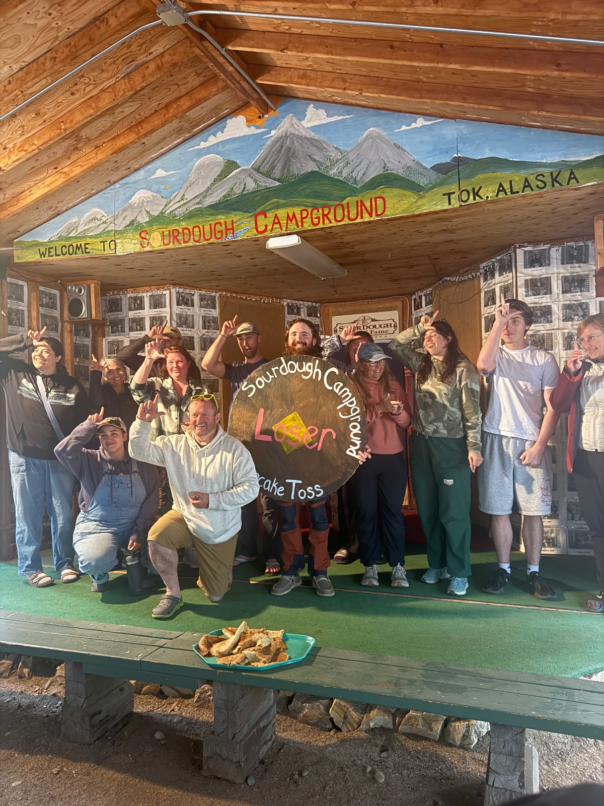 Group of visitors celebrating at Sourdough Campground in Tok, Alaska, under a log structure with mountain mural and a sign for the lodge and cake toss activity