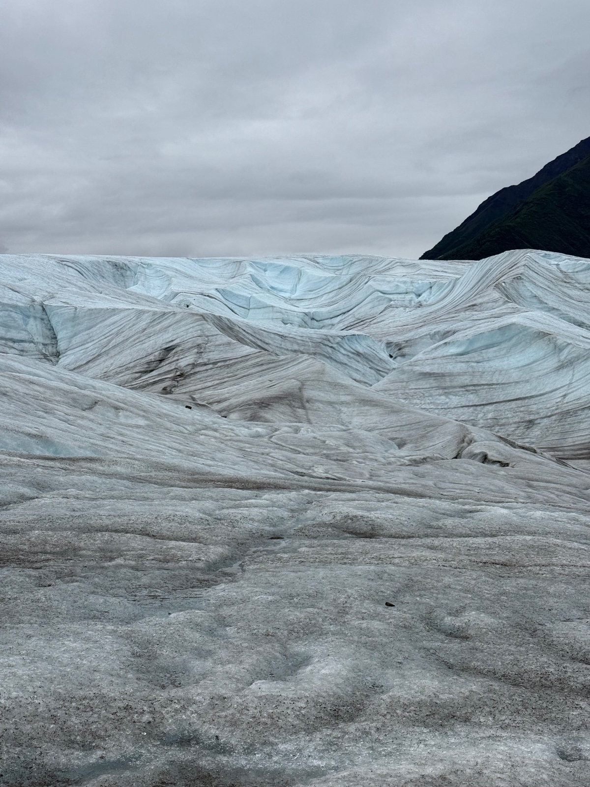 Expansive glacier with blue ice formations and dark sediment stripes, overcast sky, dark mountain visible on right side