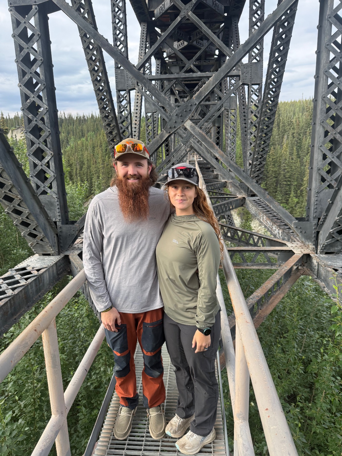 Two people wearing safety caps stand on a metal railway trestle bridge high above a coniferous forest