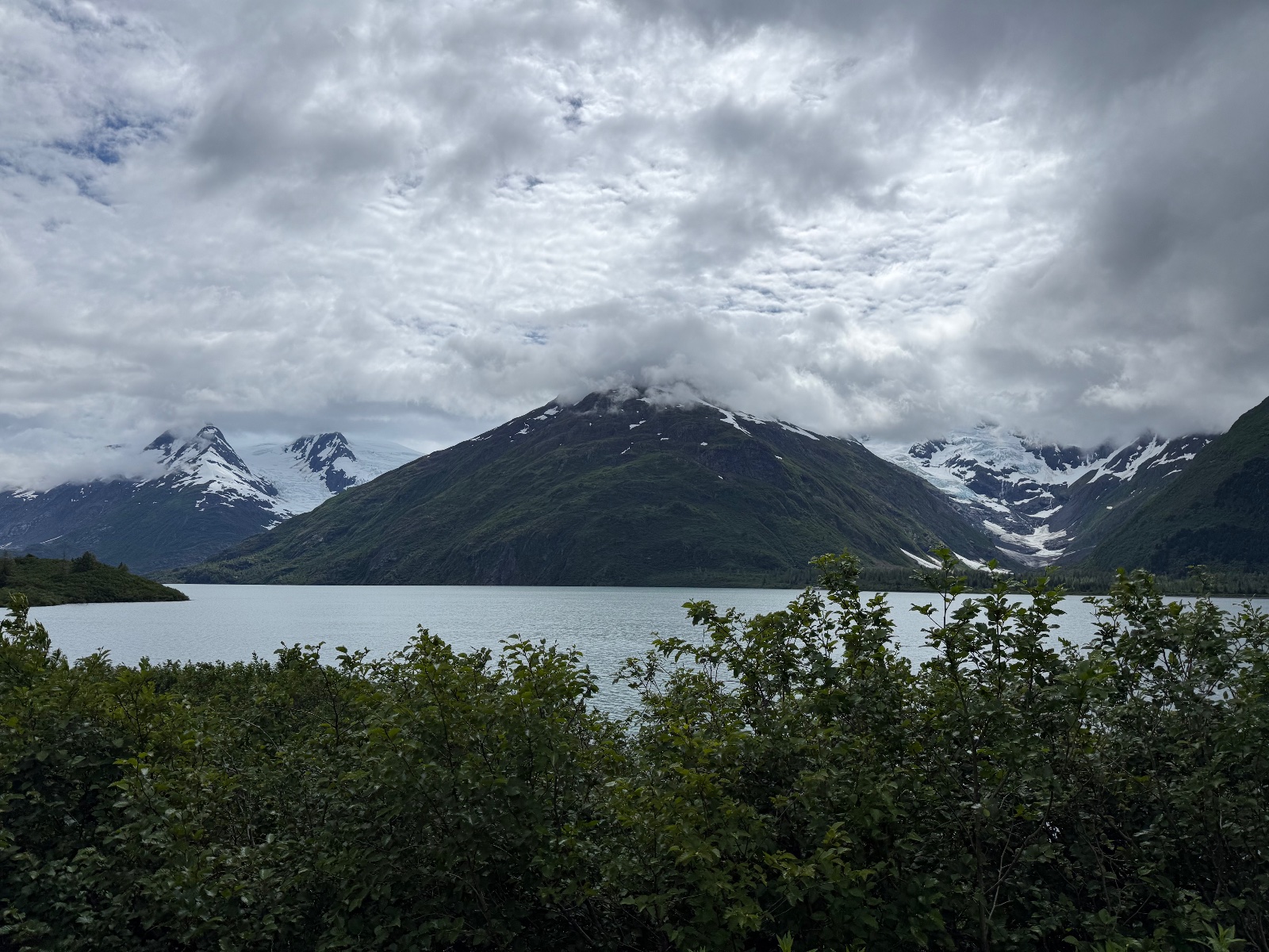 Snow-capped volcanic mountain reflected in calm alpine lake surrounded by green shrubland under dramatic cloudy sky in Alaska