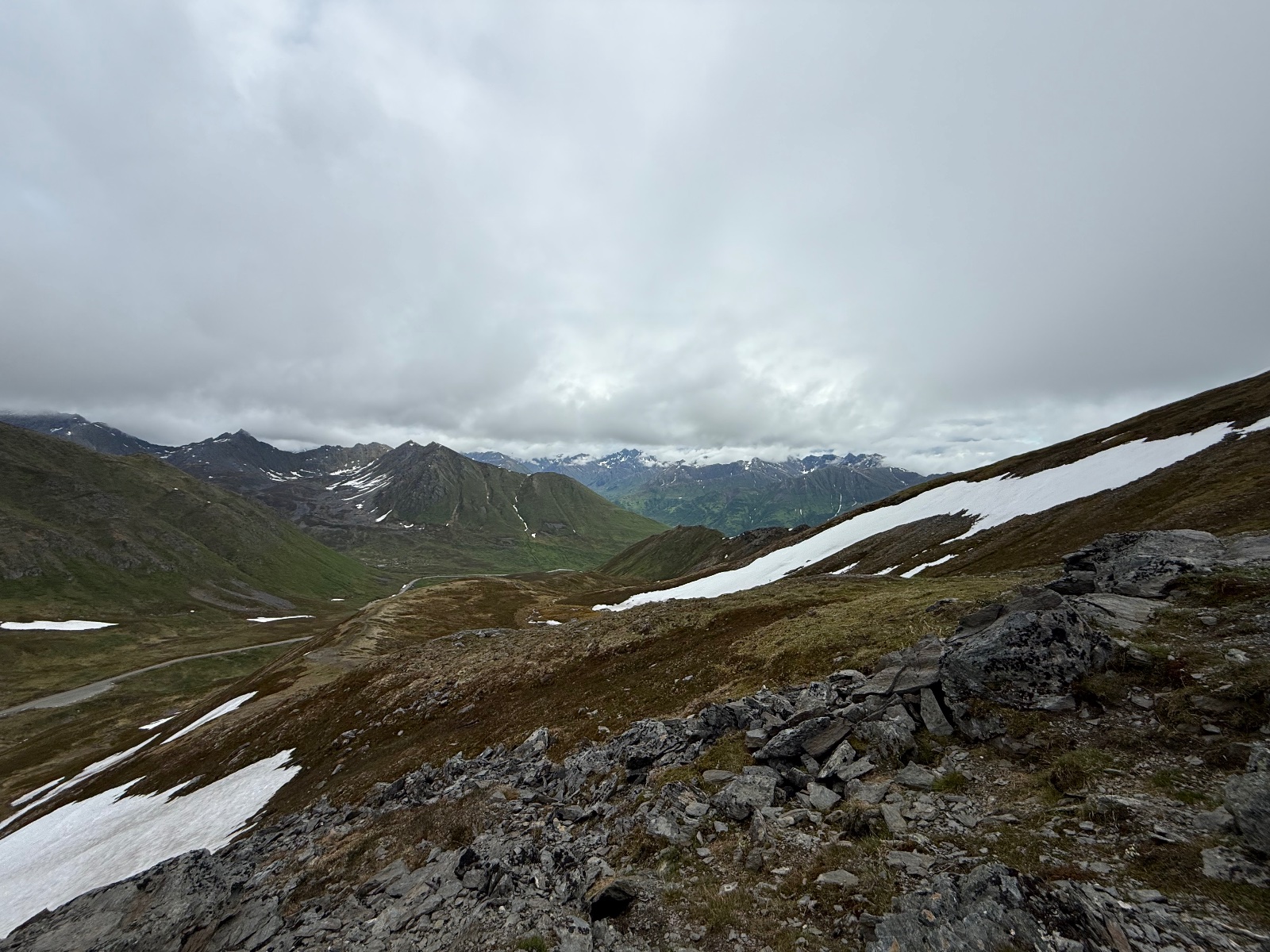 Alpine mountain valley with snow patches, rocky terrain, and distant snow-capped peaks under cloudy skies in a high-altitude wilderness area