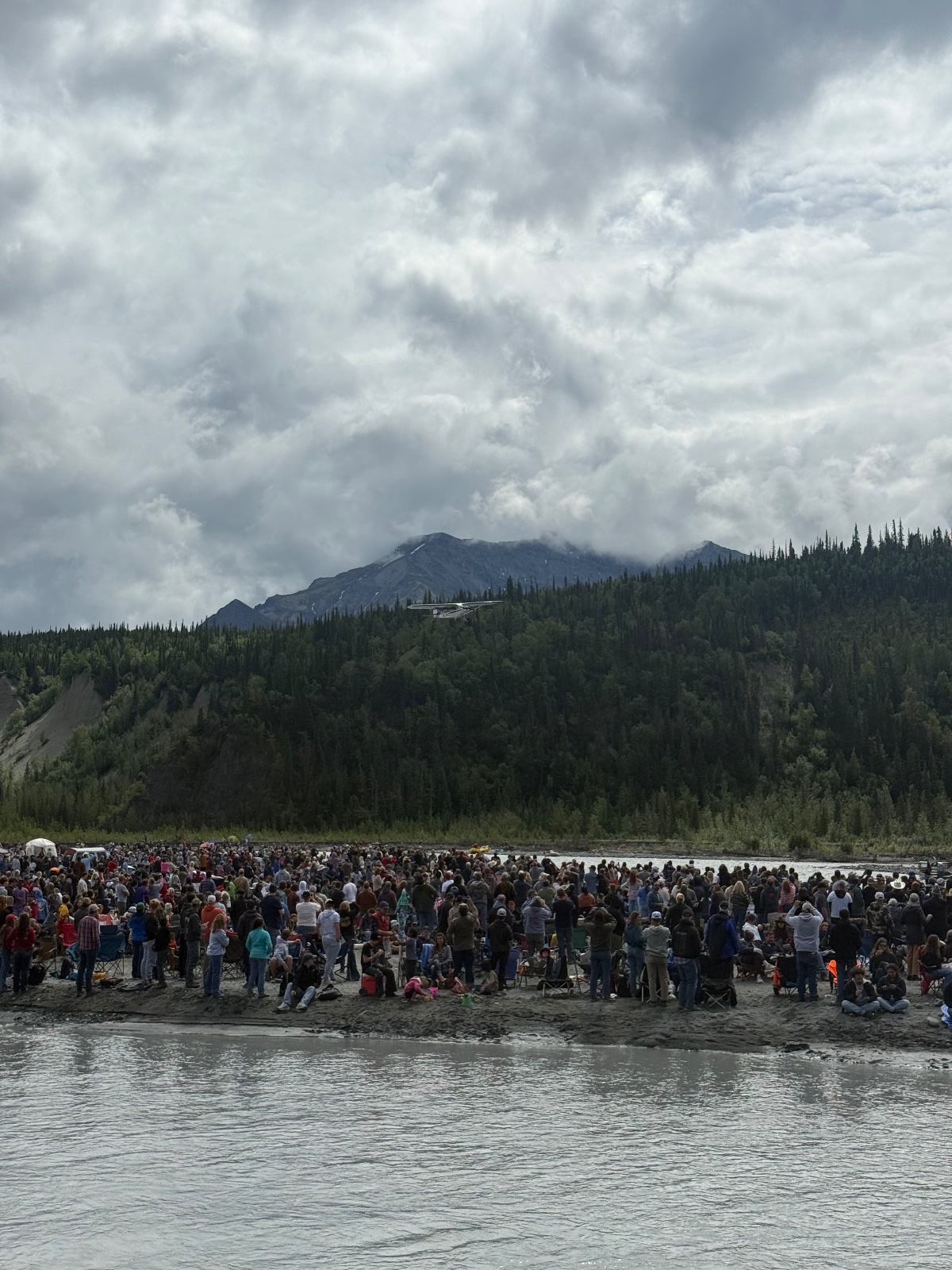 Large crowd gathered on a gravel beach along a braided river with coniferous forest and mountain backdrop under dramatic cloudy sky
