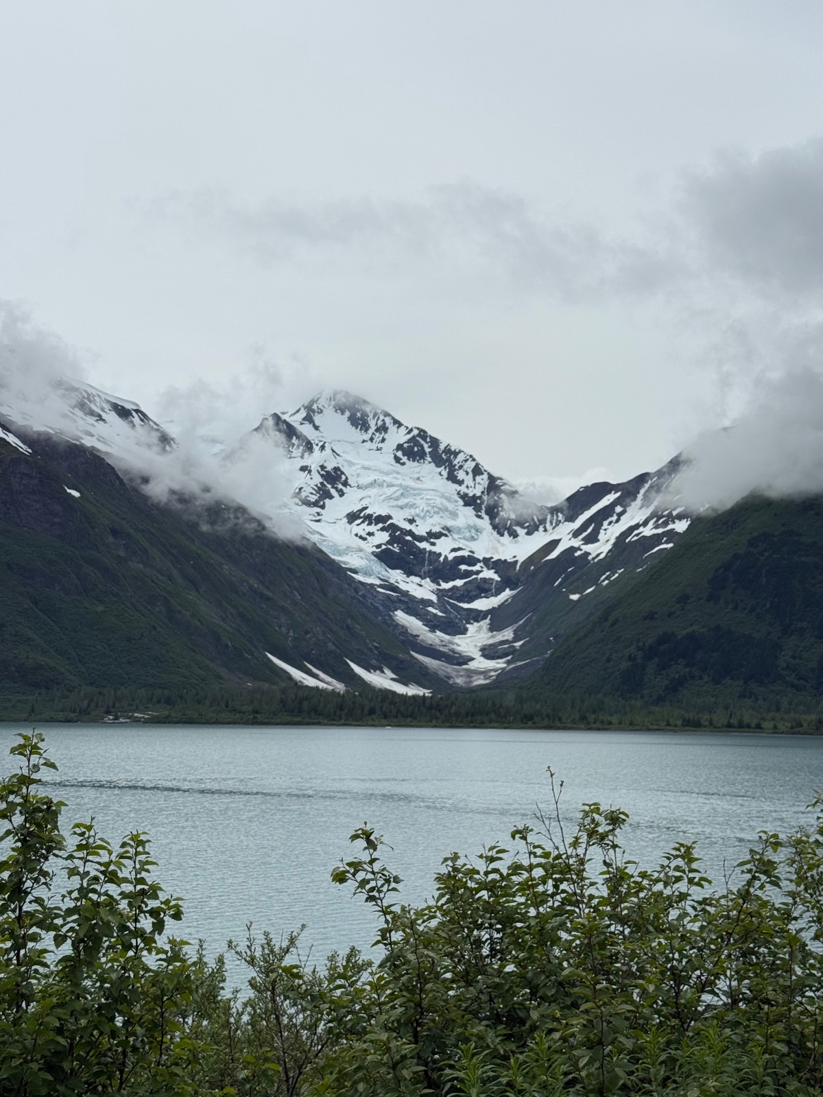 Snow-capped mountain with glacier overlooking a turquoise alpine lake surrounded by dense evergreen forest and low clouds