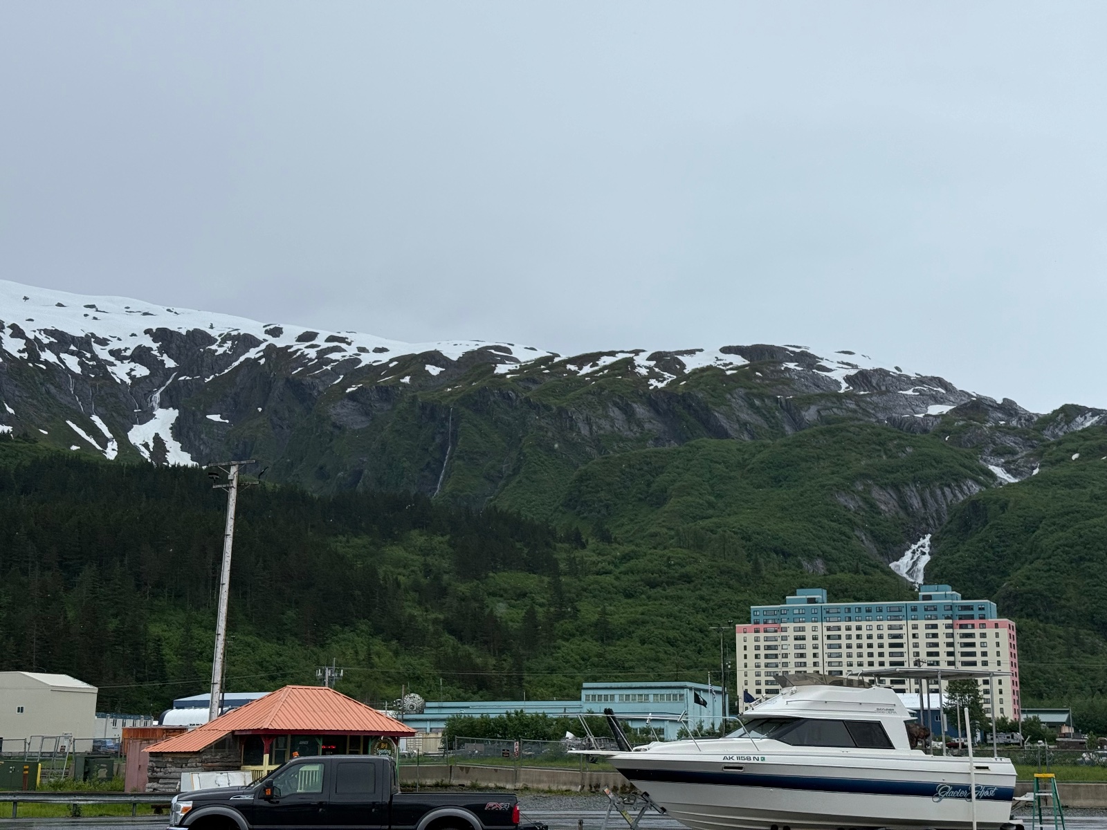 Black Ford pickup truck parked at a marina with a white cabin cruiser boat, snow-capped mountains and forested slopes visible in background under overcast sky