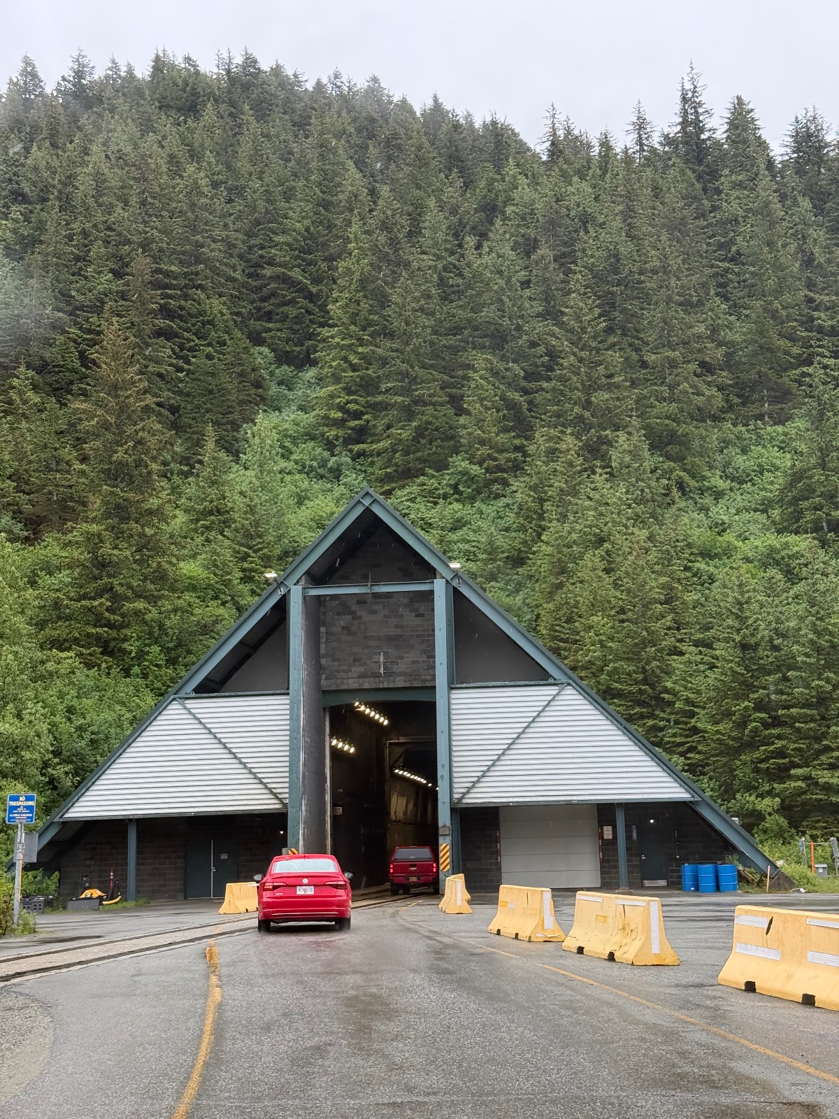 Red sedan parked in front of a modern A-frame building with yellow barriers at the entrance, surrounded by dense coniferous forest