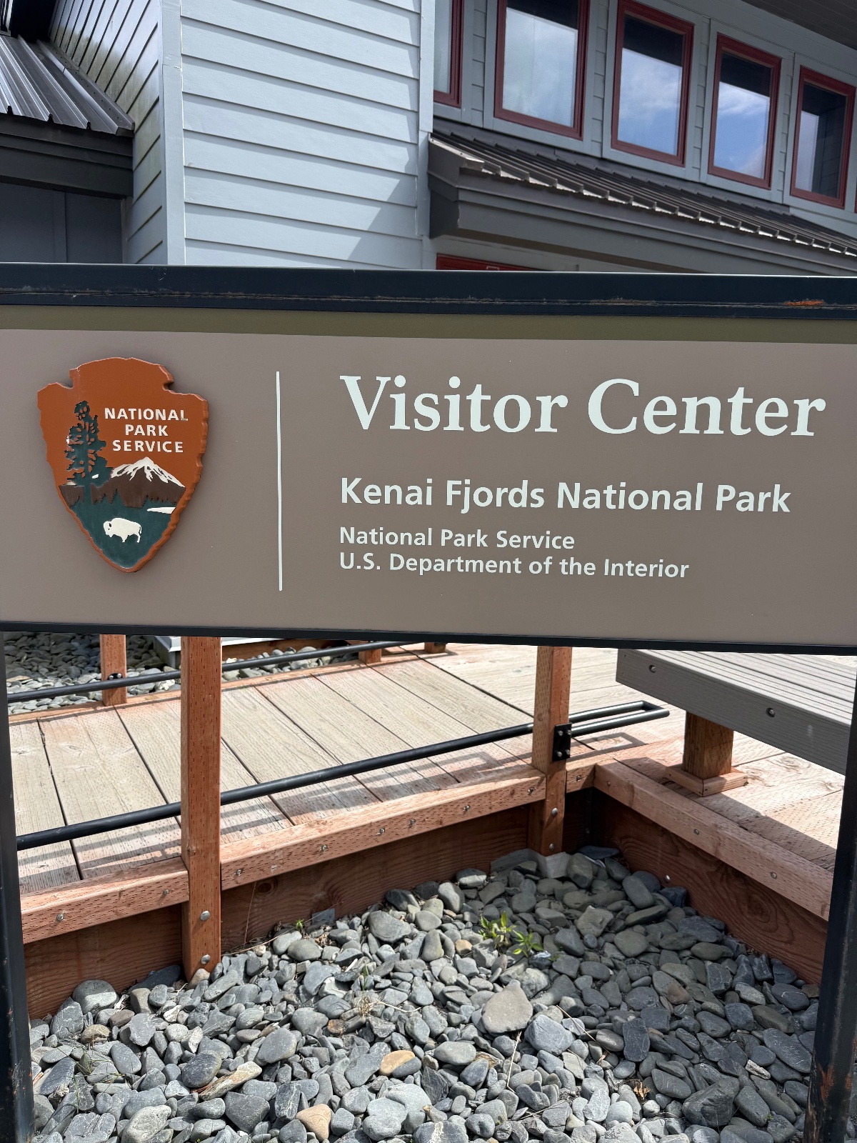 Kenai Fjords National Park Visitor Center sign with National Park Service logo mounted on tan building exterior with wooden deck and gravel landscaping