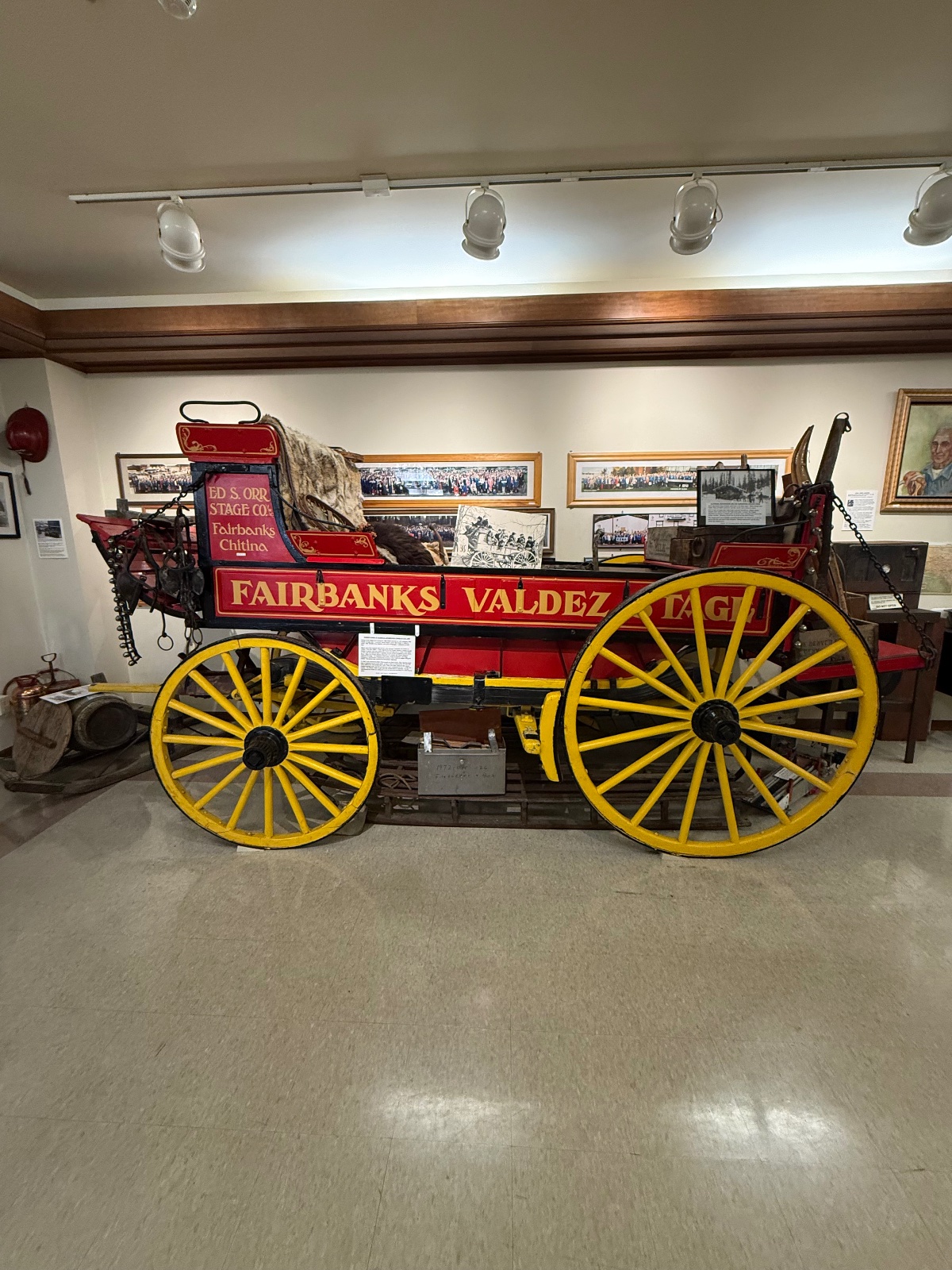 Historic Fairbanks Valdez stage coach with red body and bright yellow wheels displayed in a museum with historical photographs on the walls behind it.