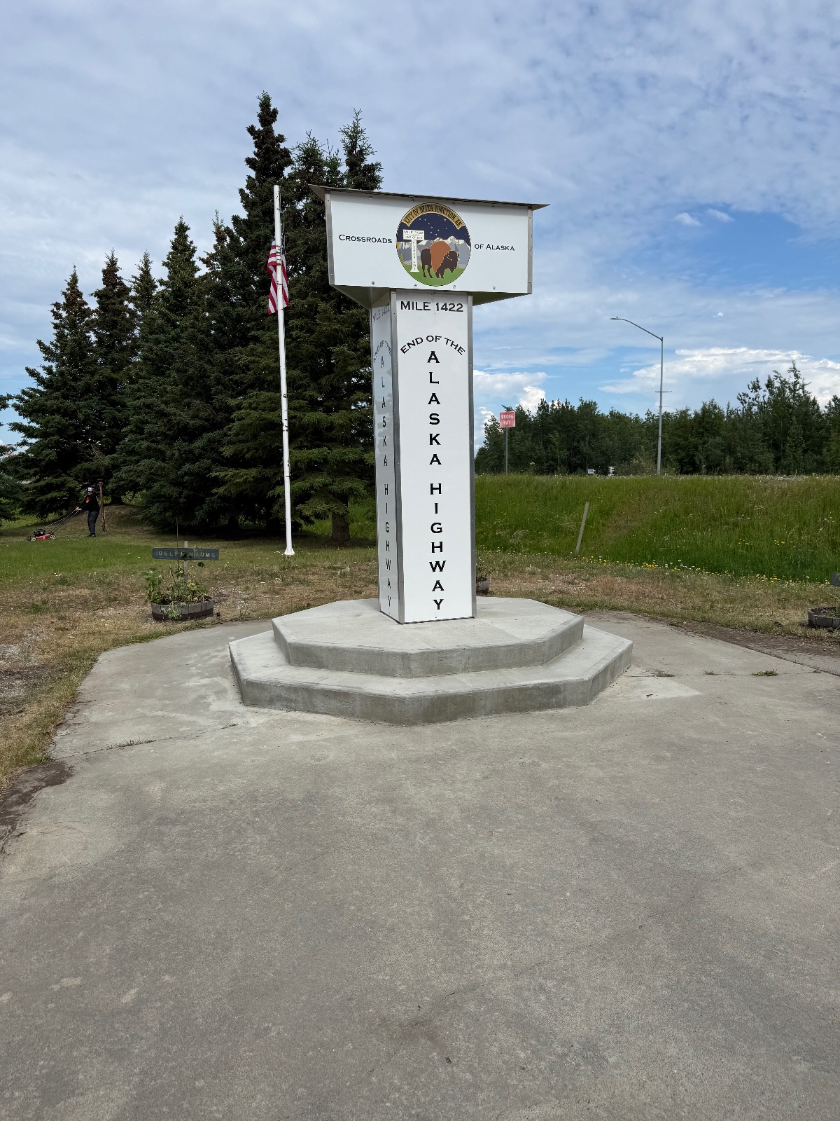 End of Alaska Highway milestone marker at Mile 1422 in Crossroads of Alaska, with conifer trees and grassy landscape under partly cloudy sky