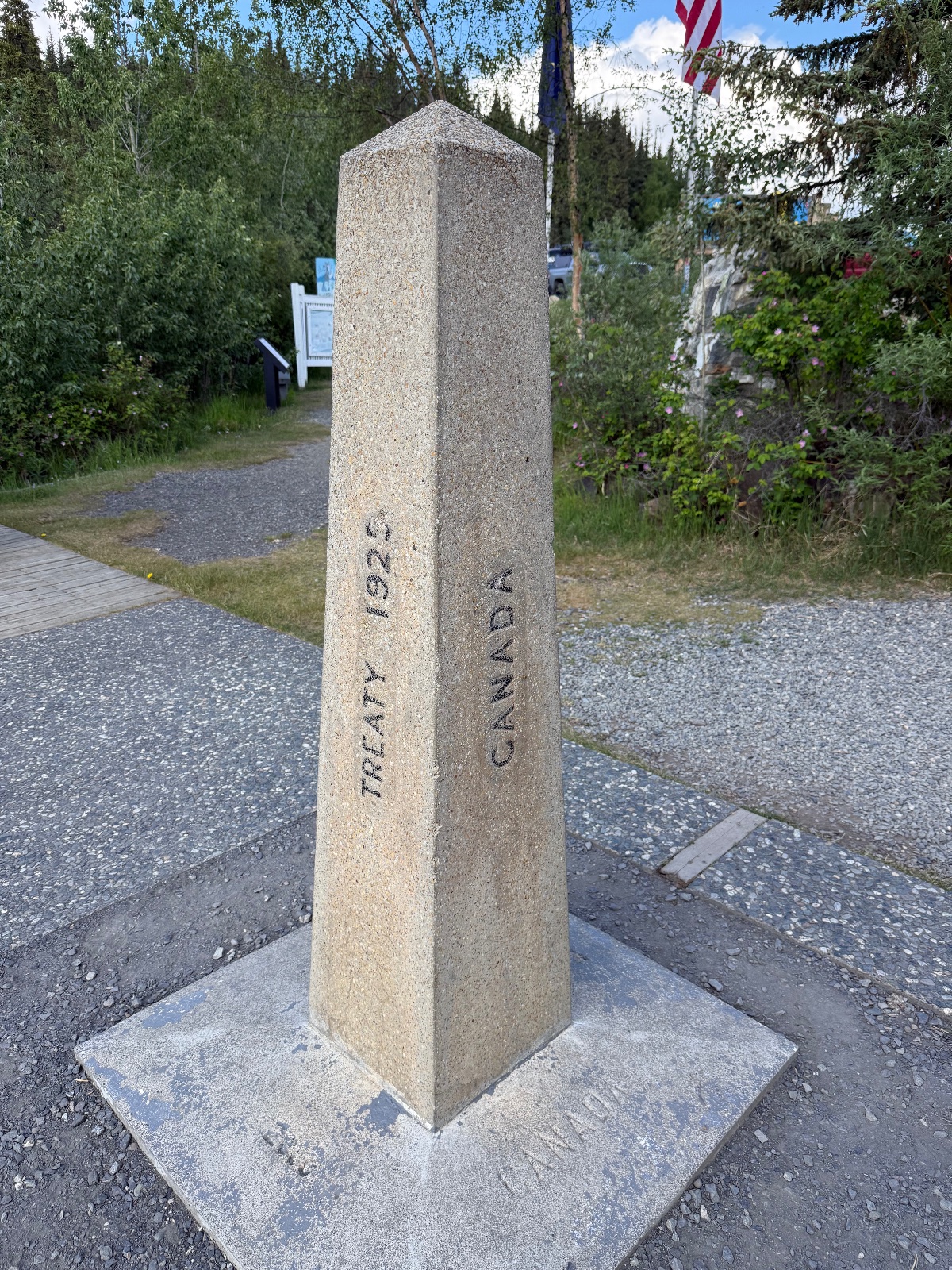 Stone monument marker engraved with 'TREATY 1921' and 'CANADA' at a scenic overlook with mountains, forest, and American flag visible in background