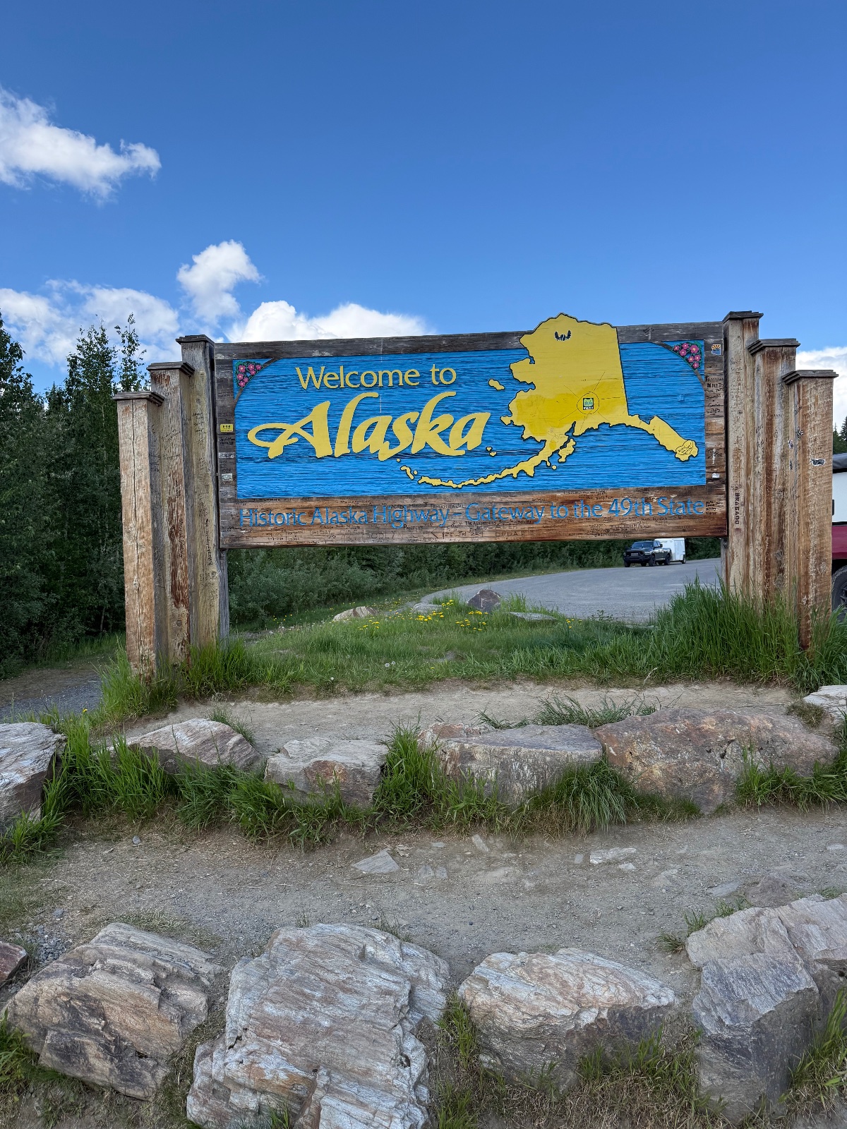 Welcome to Alaska sign with yellow state outline on blue wooden board, roadside landmark with evergreen trees and clear blue sky