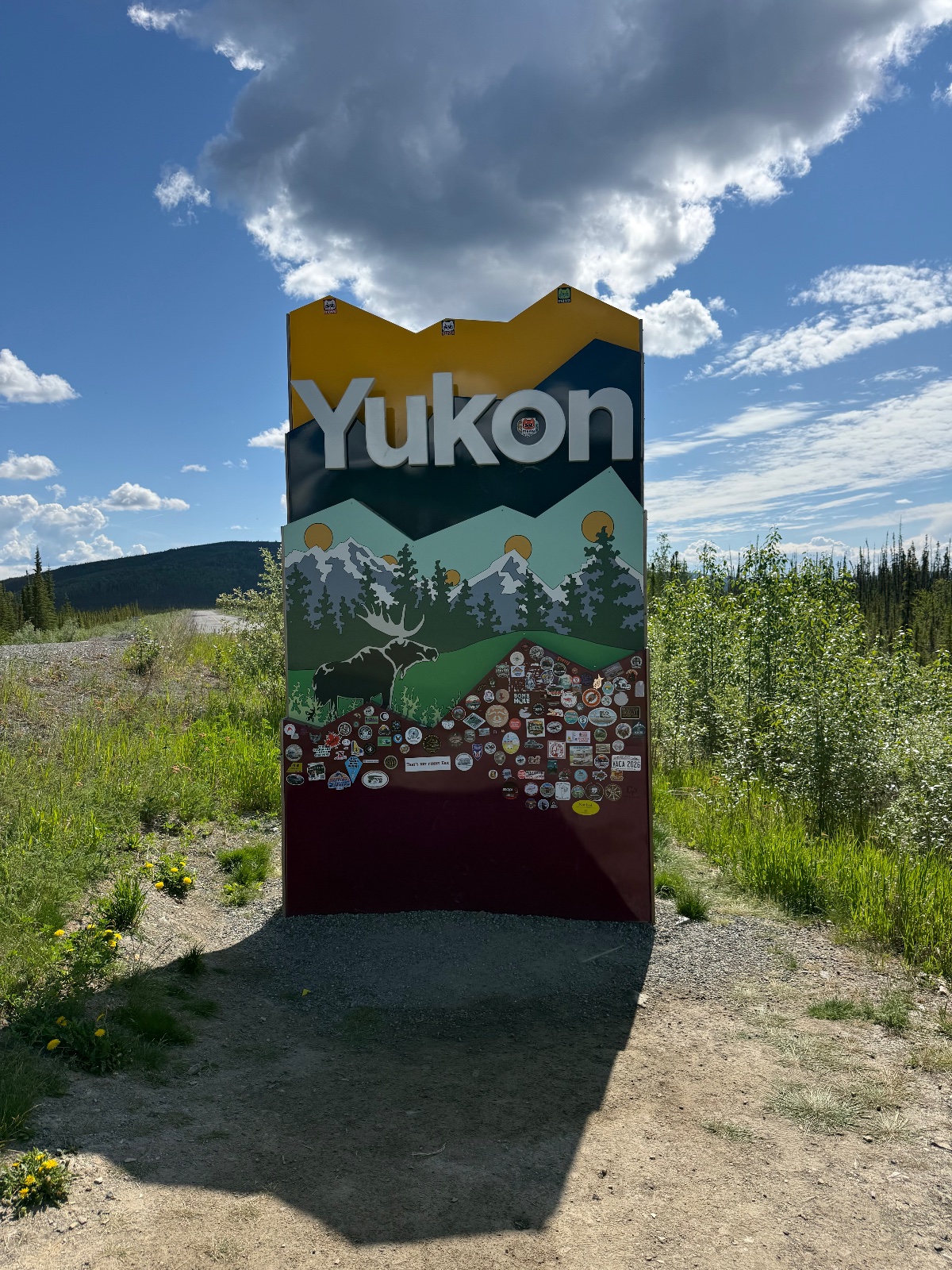 Yukon welcome sign with colorful visitor patches on a gravel road surrounded by boreal forest and mountains under partly cloudy sky
