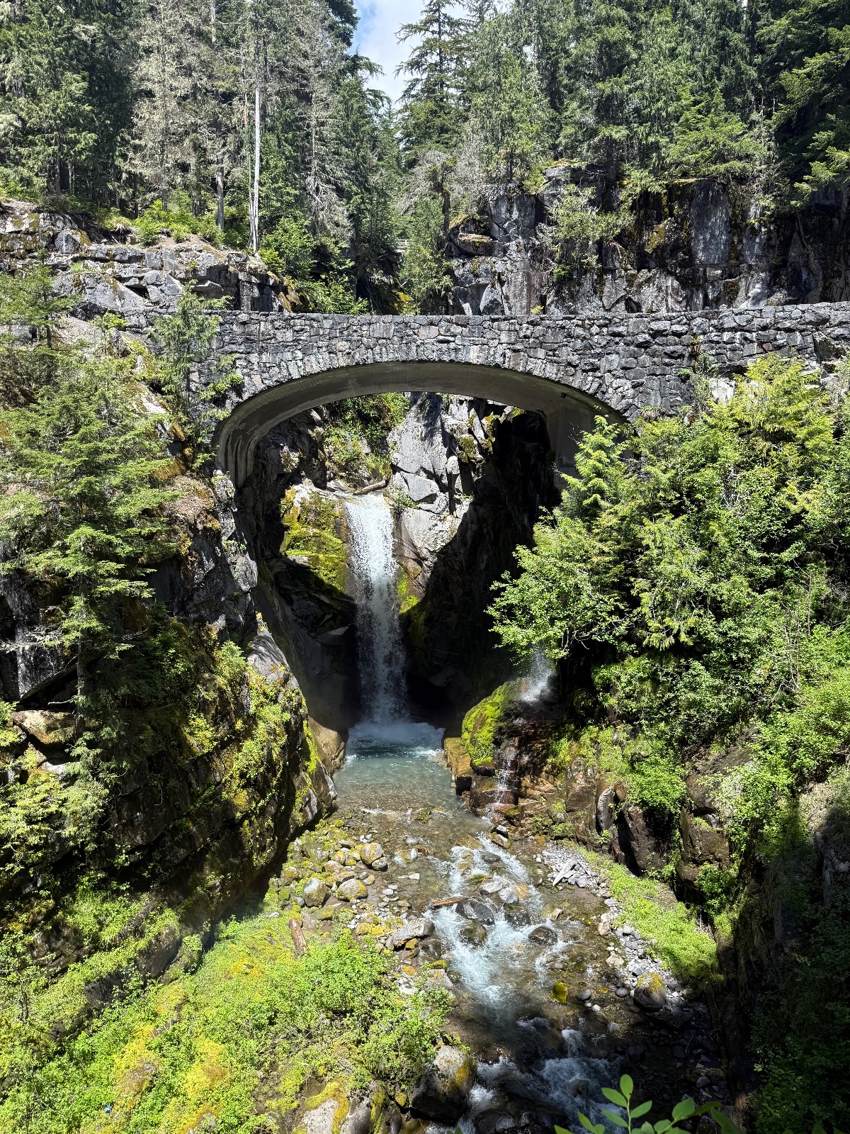 Stone arch bridge spanning a mountain stream with waterfall in lush forest canyon surrounded by tall evergreen trees and moss-covered rocks