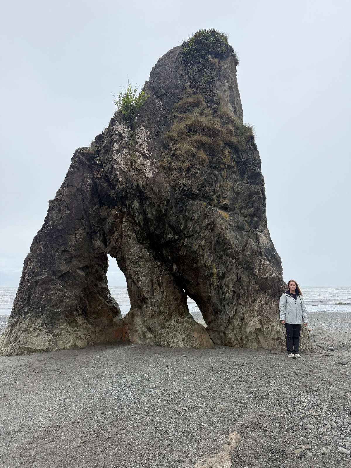 Person standing in front of a massive natural rock arch formation on a sandy beach with ocean and overcast sky in background