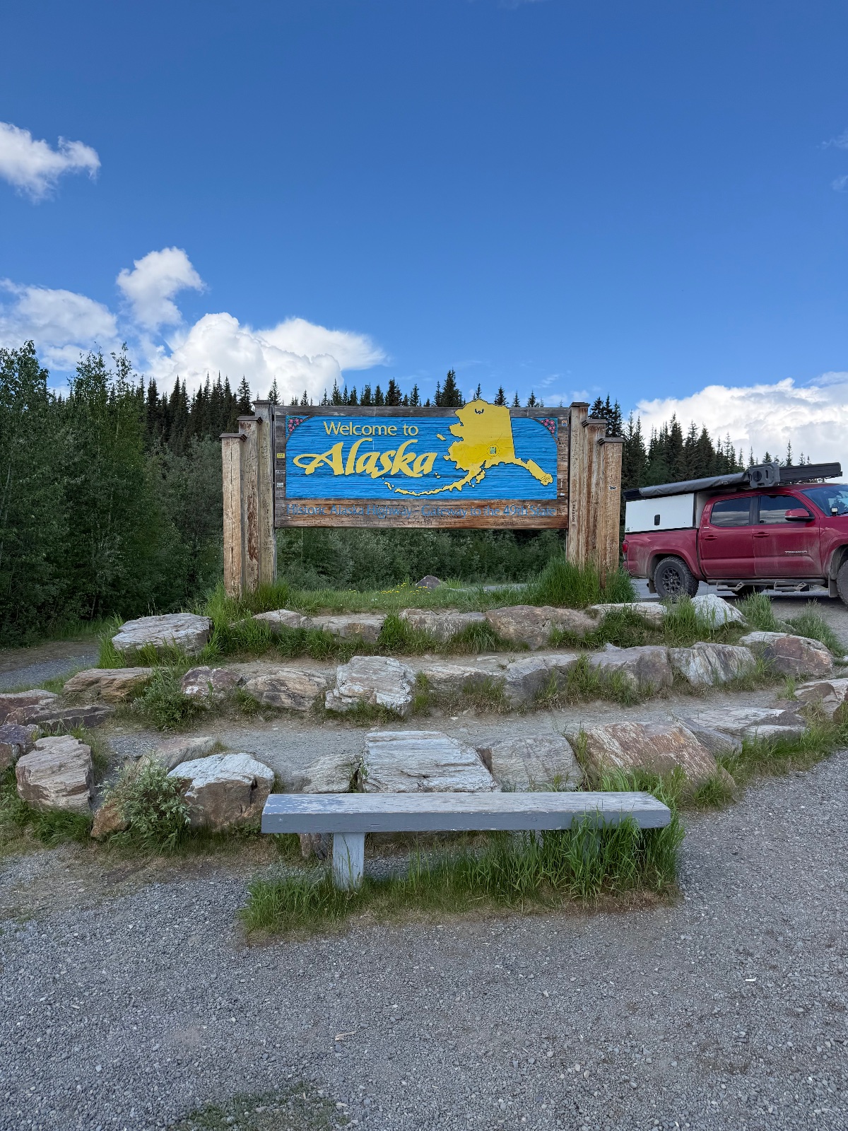 Welcome to Alaska sign at a rest area with a red pickup truck, wooden bench seating, and evergreen forest under blue sky