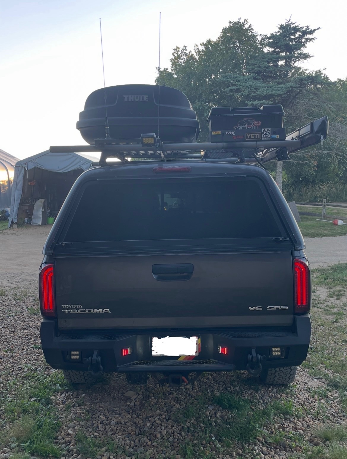 Black Toyota Tacoma V6 with Thule roof rack and cargo boxes parked at a campground with trees in background