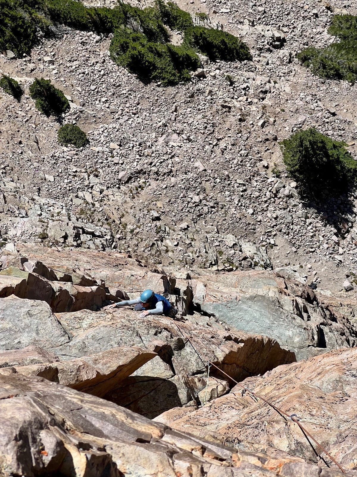 Rock climber in blue helmet rappelling down a steep granite cliff face with loose scree slope and desert vegetation above