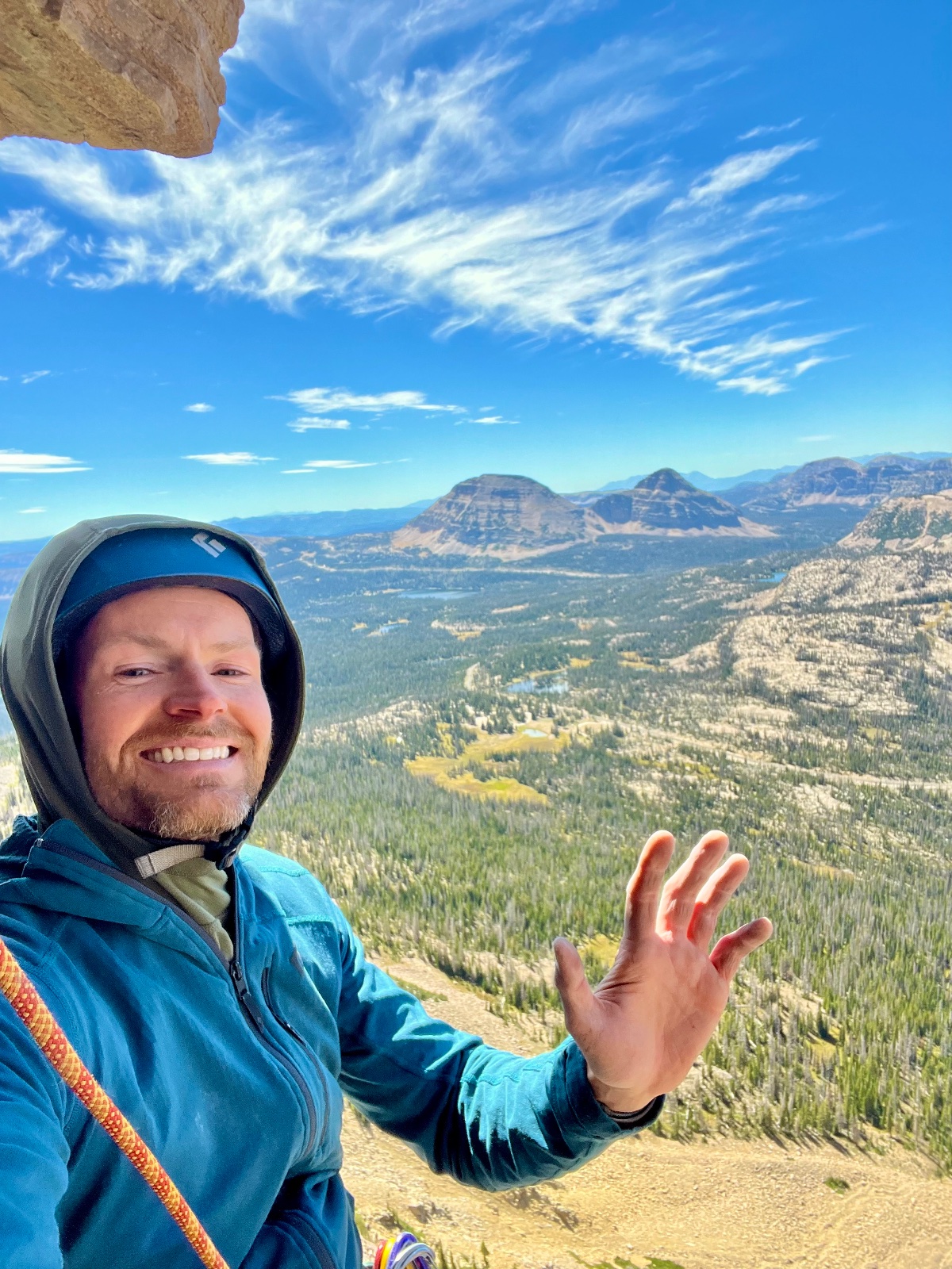 Smiling hiker in blue jacket waving at high alpine overlook with forested valley, mountain peaks, and clear blue sky in background