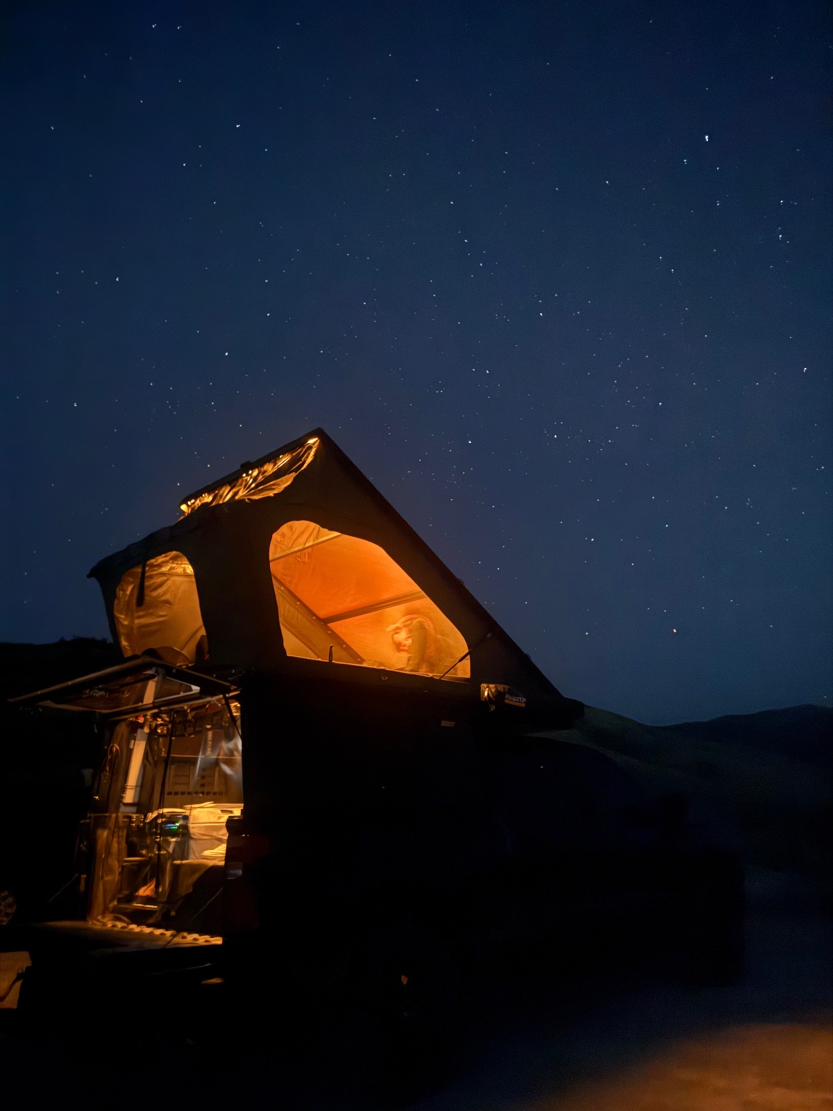 Illuminated rooftop tent on vehicle under starry night sky with mountains silhouetted in background