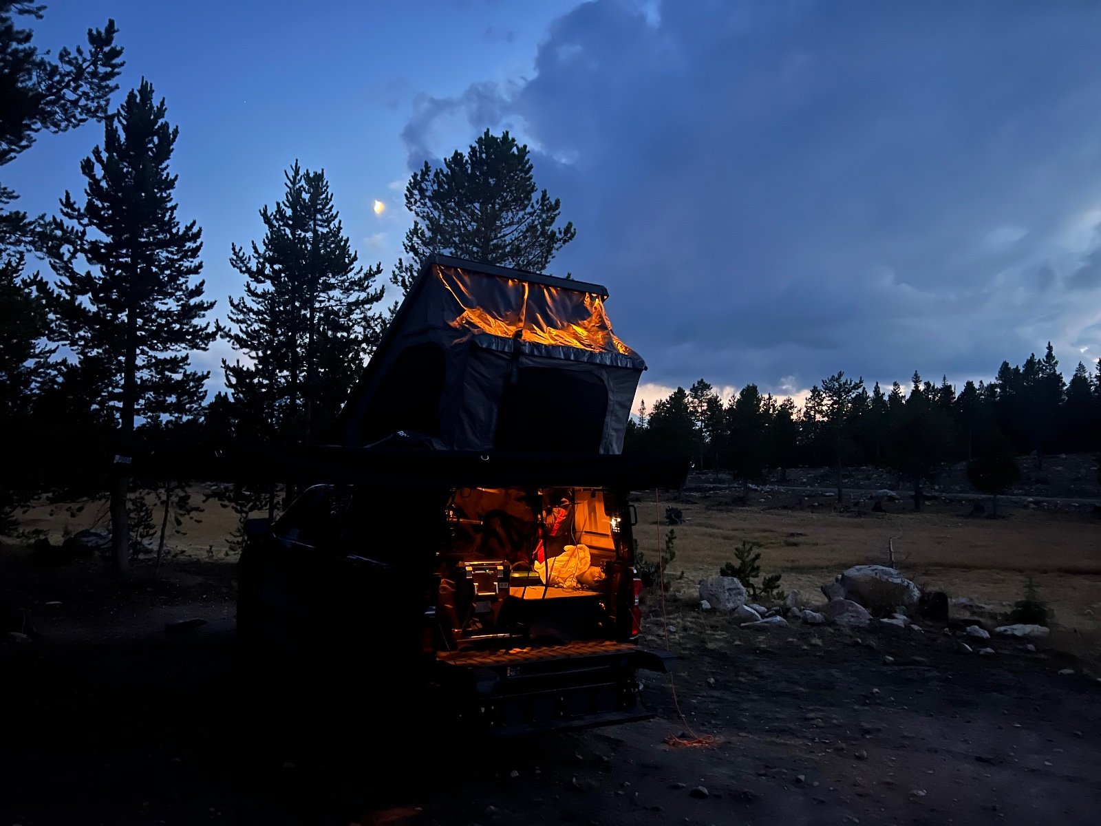 Illuminated van with pop-up roof camper setup in a pine forest clearing at dusk with moon visible in twilight sky
