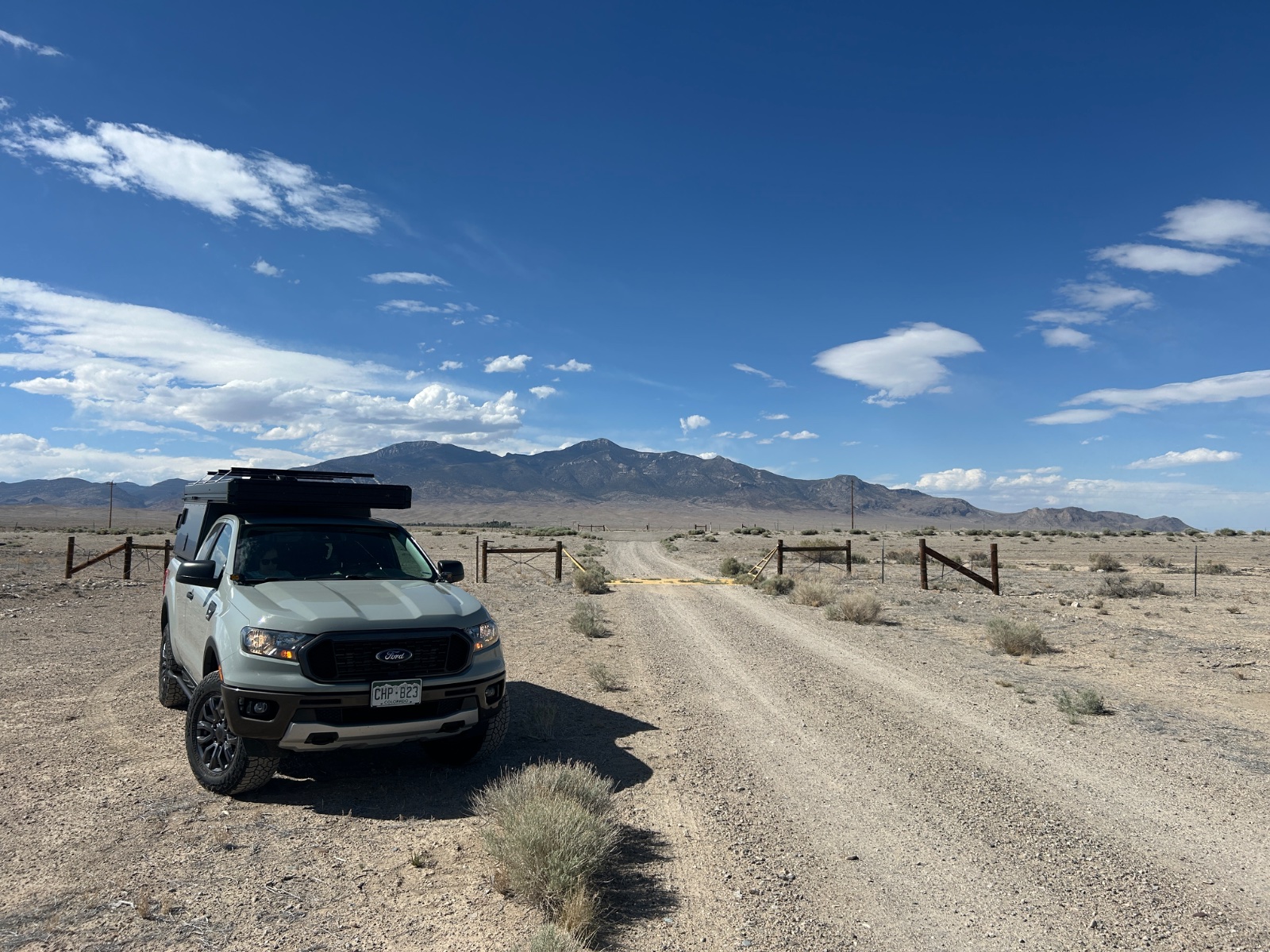 White Ford Ranger with roof rack parked on desert dirt road near wooden ranch fence with mountains in background under blue sky