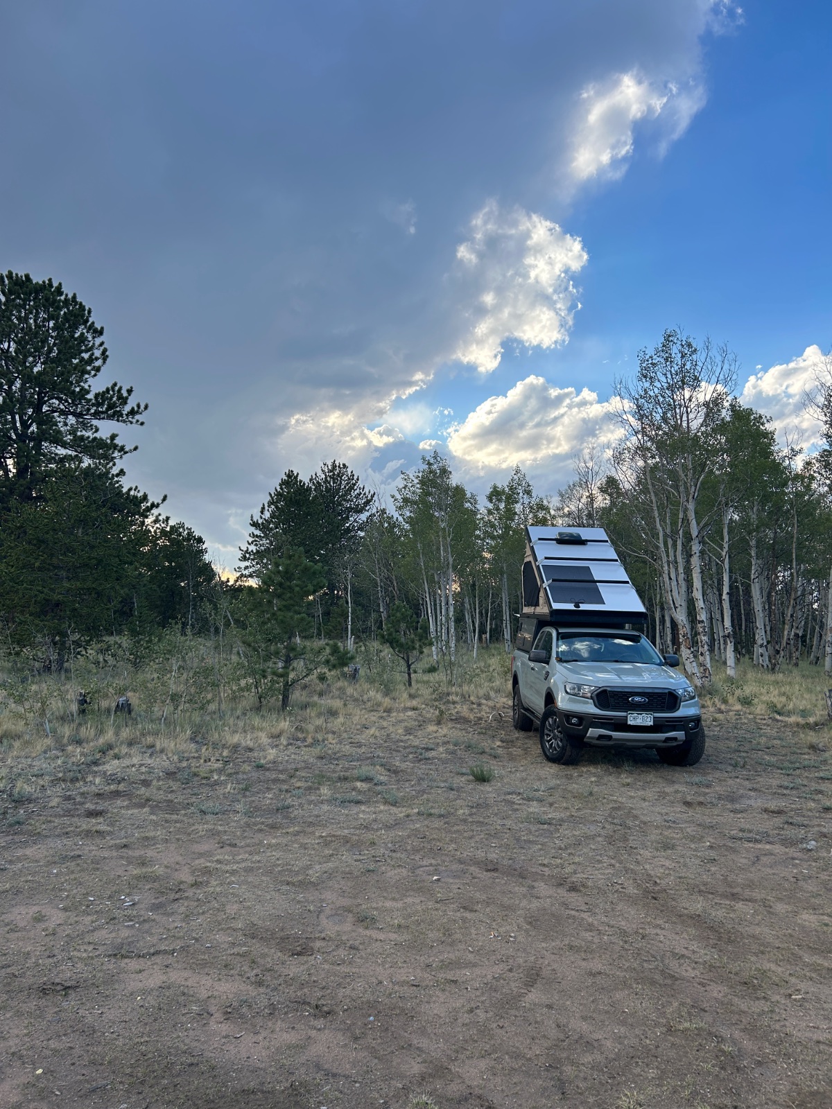 White Ford Ranger with roof-mounted solar panels and pop-up camper in a pine forest clearing under blue sky with white clouds