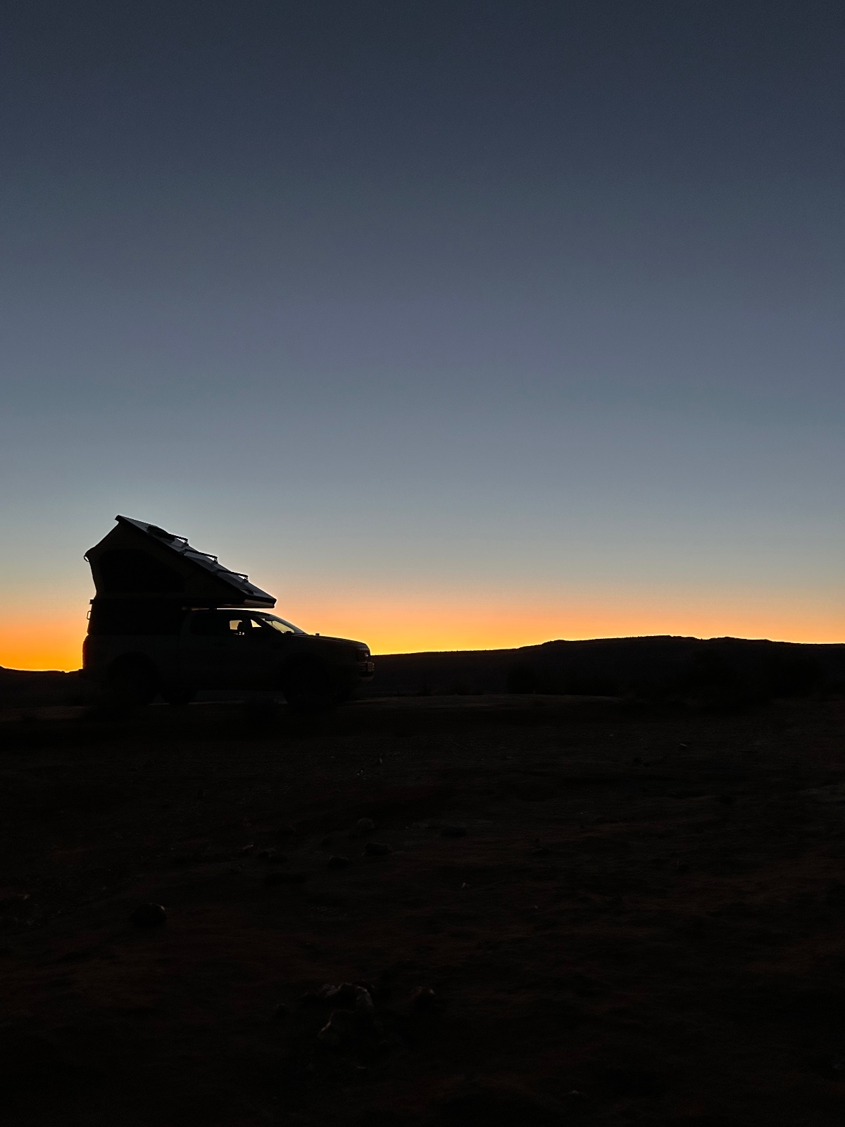 Silhouetted SUV with pop-up roof tent parked on desert terrain at golden hour sunset with mountains on horizon