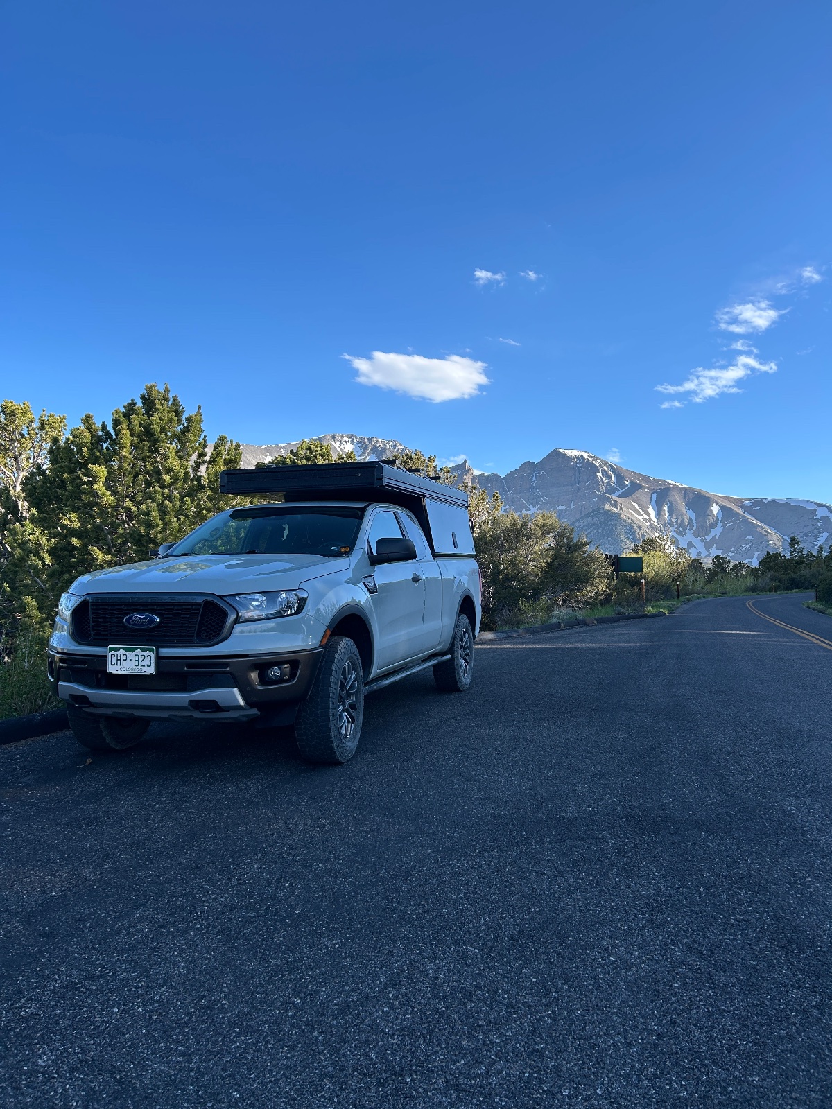 White Ford Ranger pickup truck with roof rack parked on a paved road with snow-capped mountains in the background under clear blue sky
