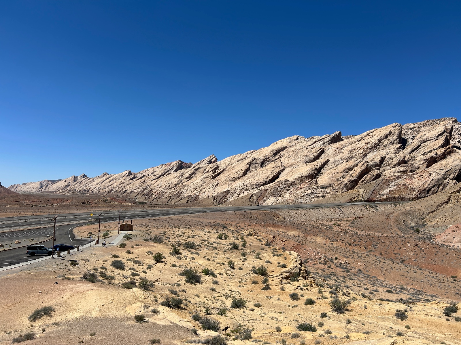 Desert landscape with layered rock formations, sparse vegetation, paved road, and parked vehicles near a rest stop structure under clear blue sky