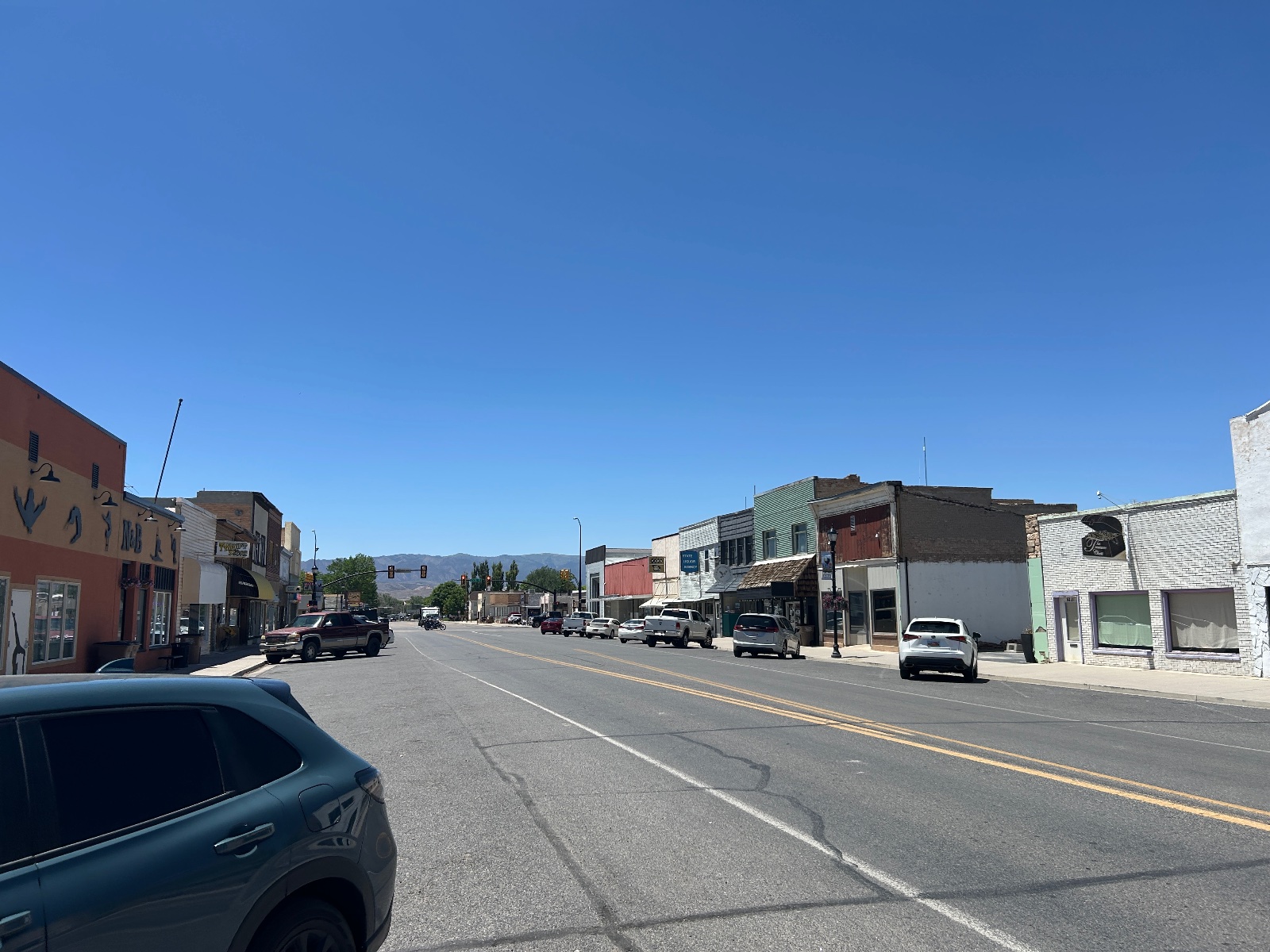Main street of a small town with vintage brick buildings, parked vehicles, mountains visible in the distance under clear blue sky