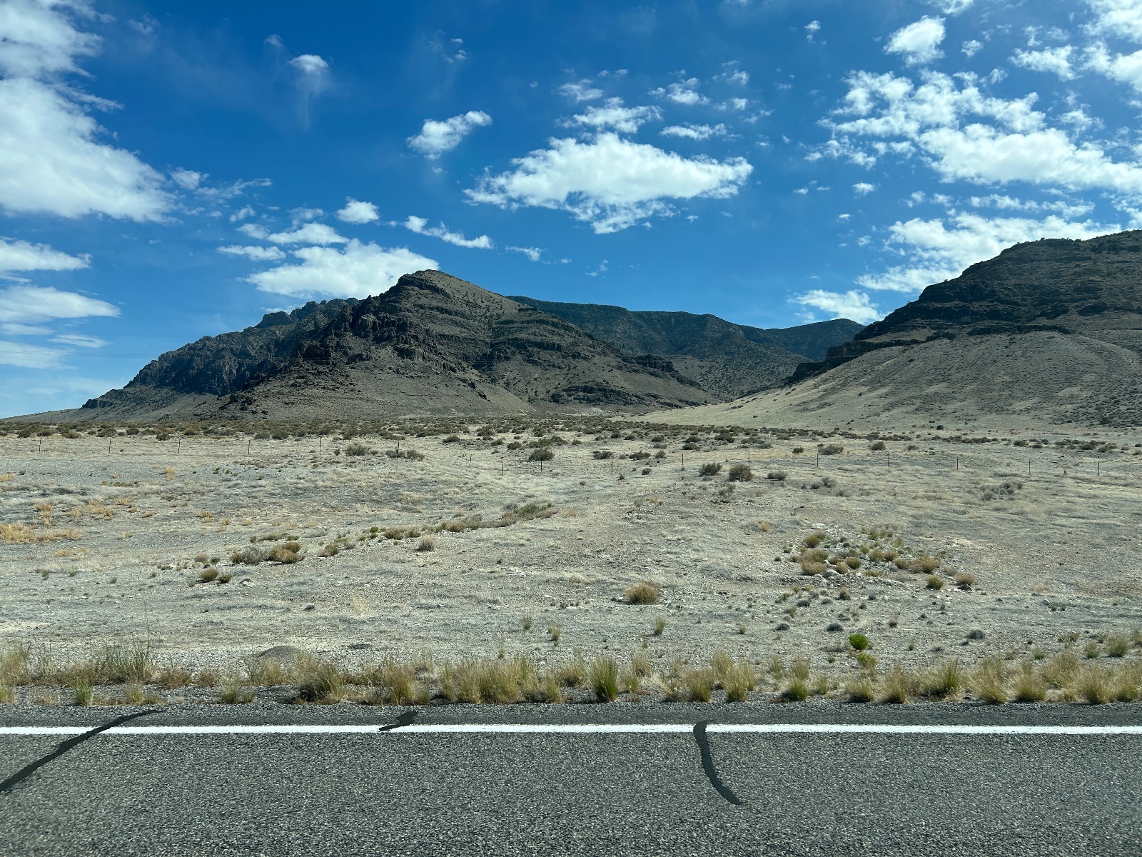 Arid high-altitude desert landscape with sparse vegetation, rocky mountains, fence line, and clear blue sky with scattered clouds