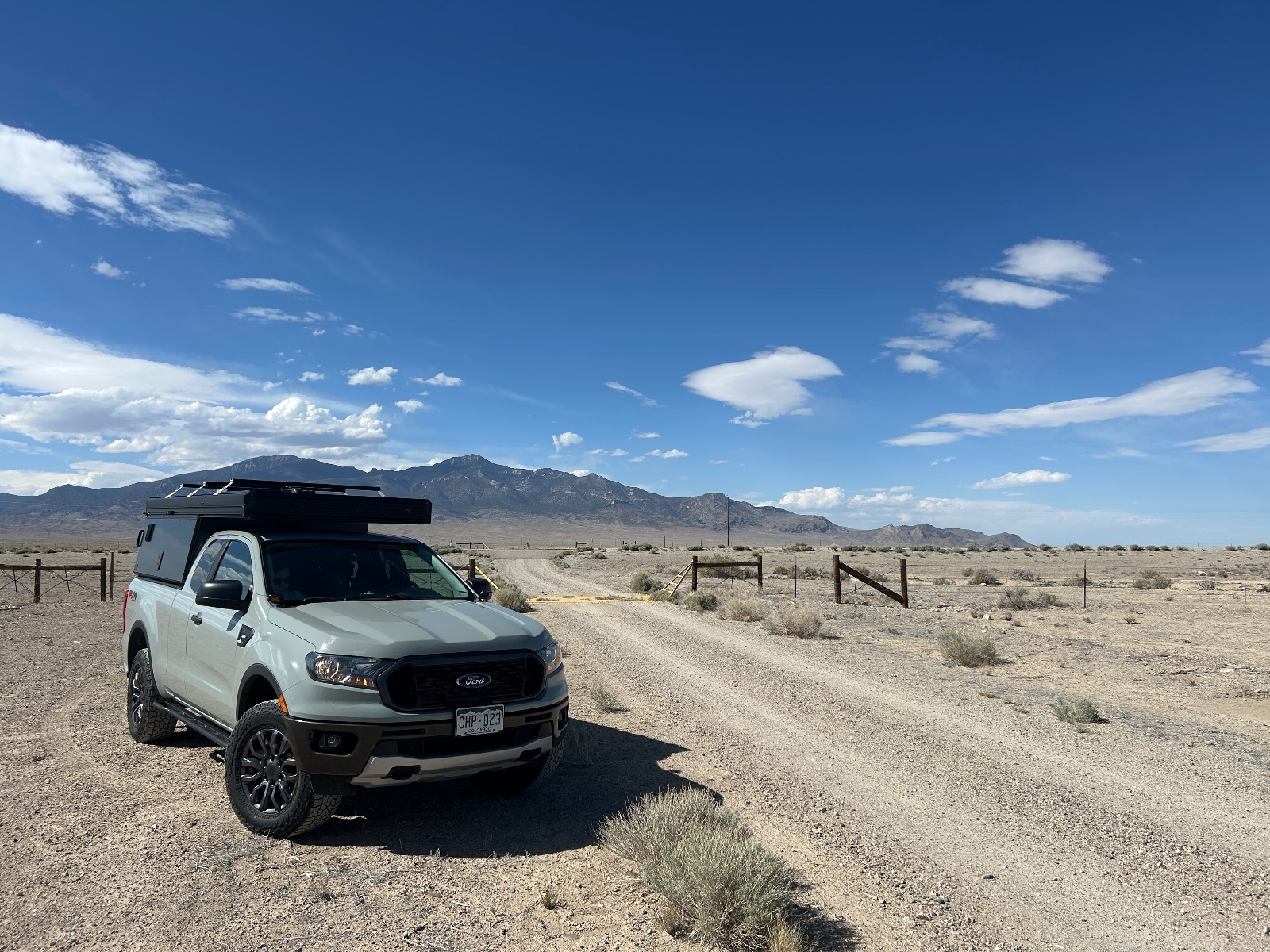 White Ford Ranger with pop-up camper parked on desert ranch road with mountains in background under blue sky
